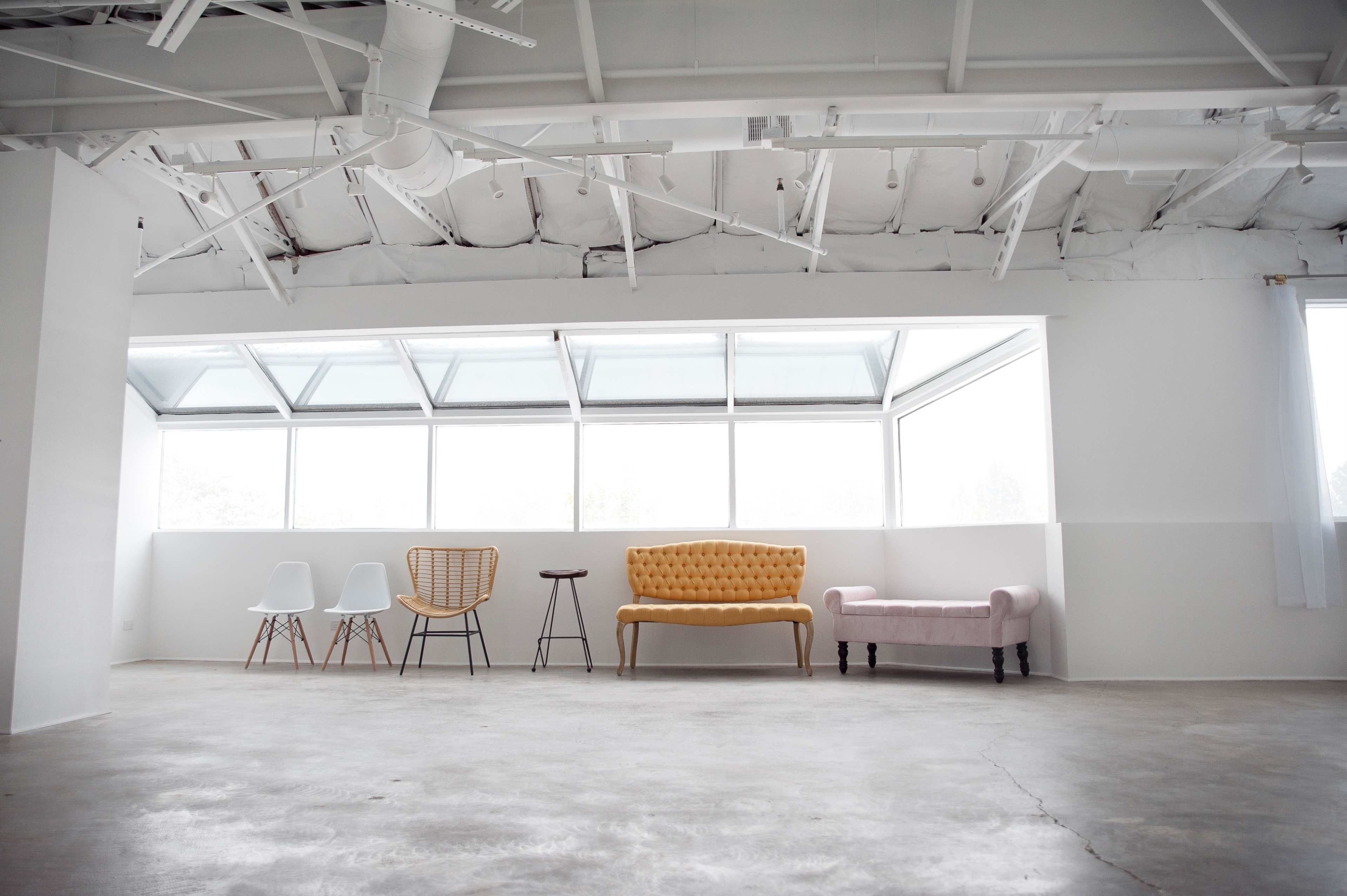 A bright, minimally furnished room with a large window, featuring a yellow sofa, a pink chair, various other seats, and a concrete floor.