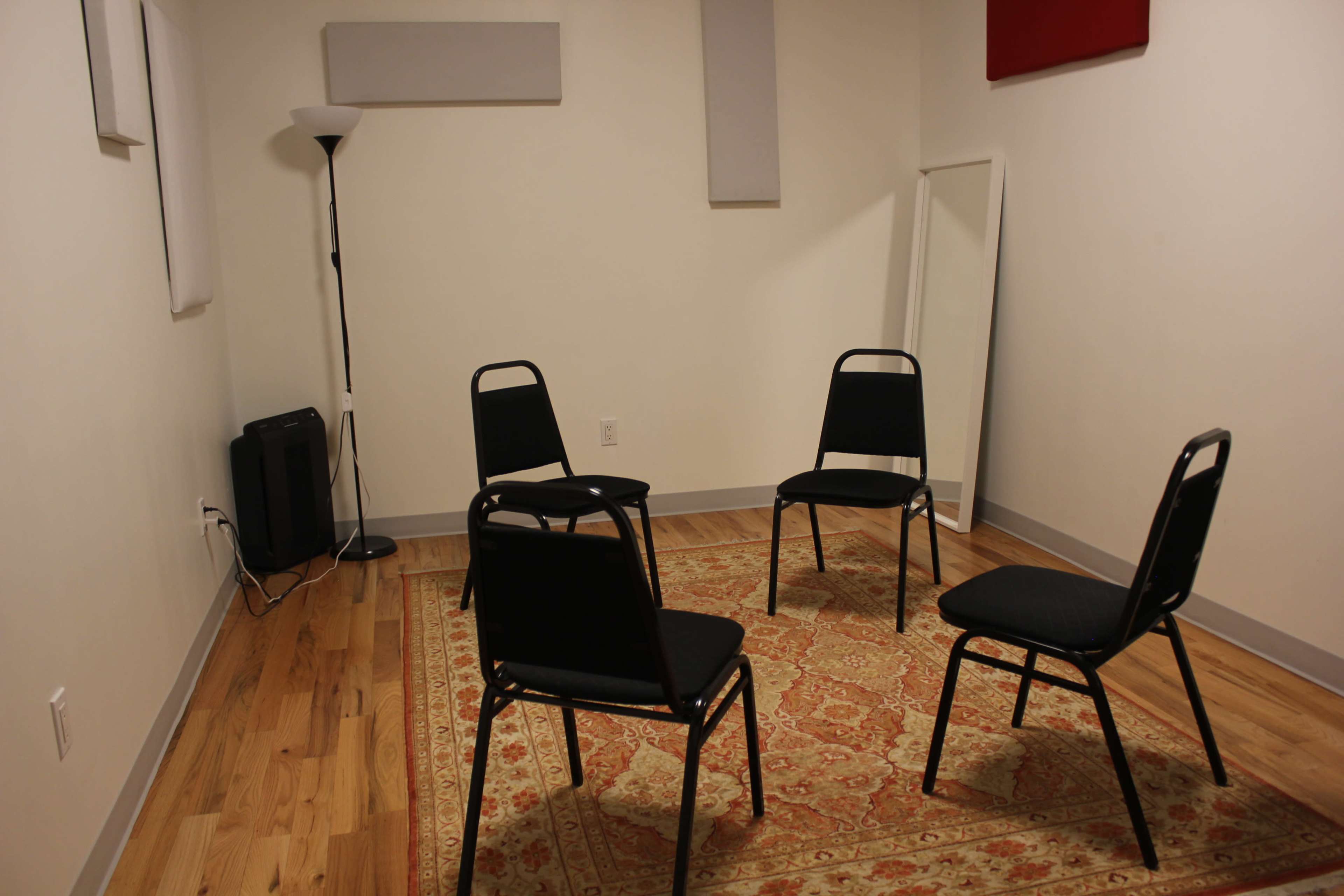 Four black chairs arranged in a square on a patterned rug in a sparsely decorated room with a floor lamp and a full-length mirror.