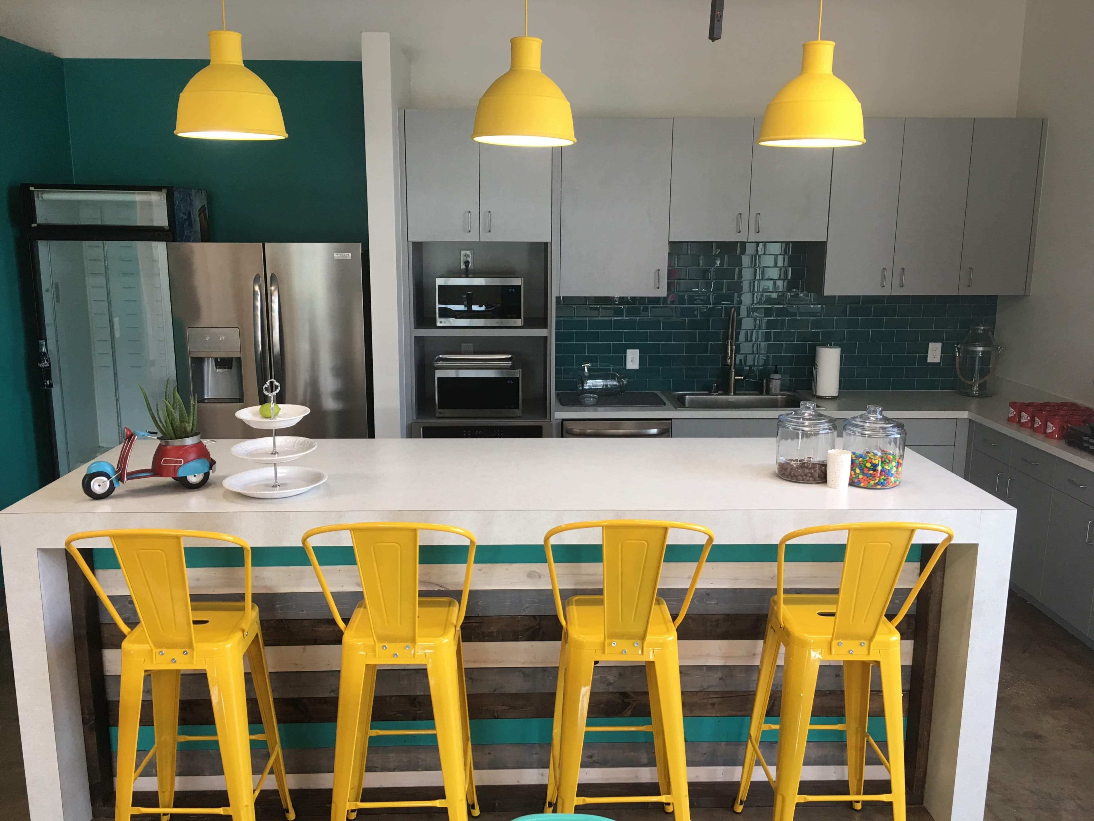A modern kitchen features a central island with four yellow stools, stainless steel appliances, and teal-accented walls.