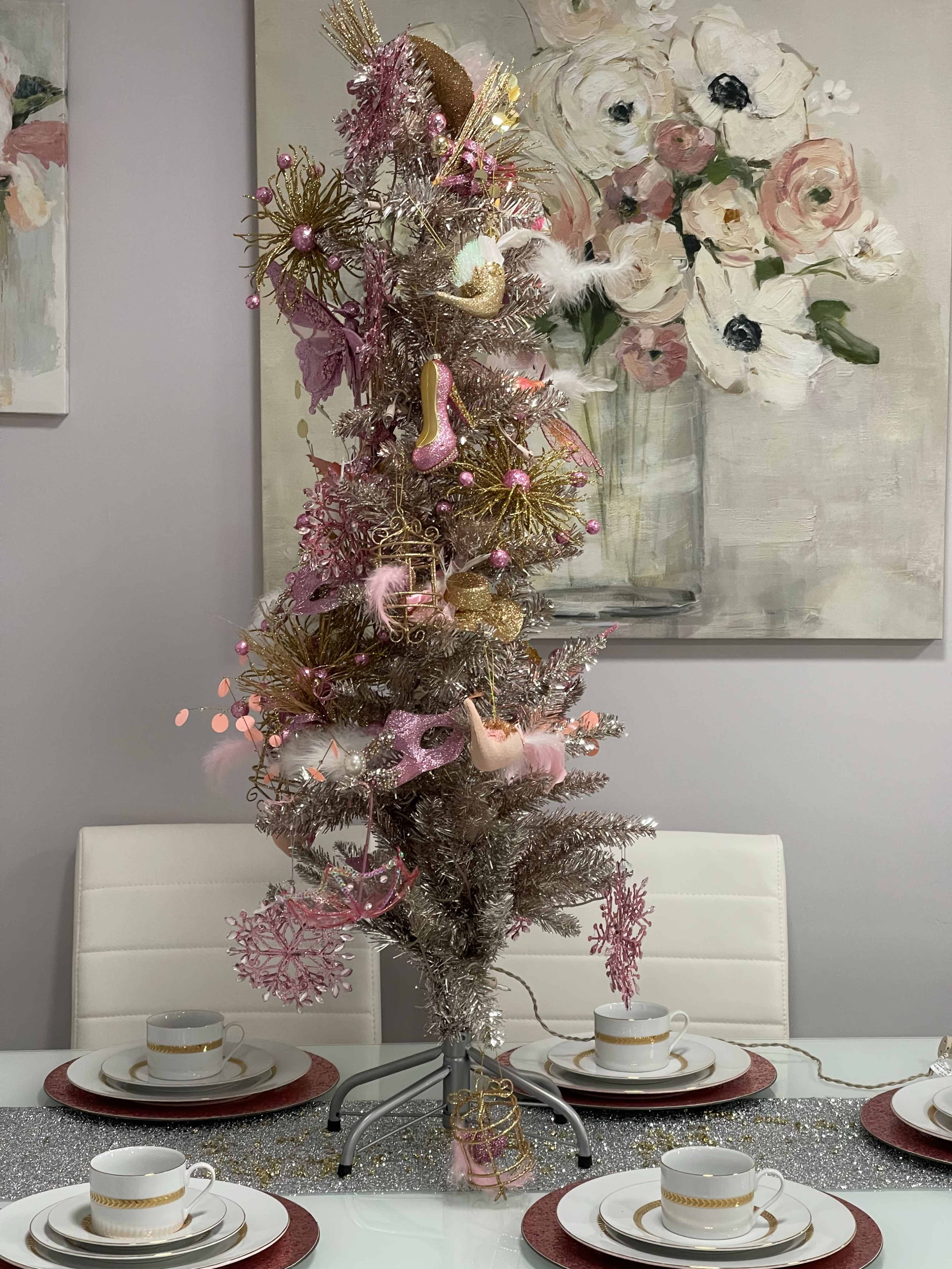 A decorative silver Christmas tree adorned with pink ornaments sits on a dining table set with white cups and red placemats.