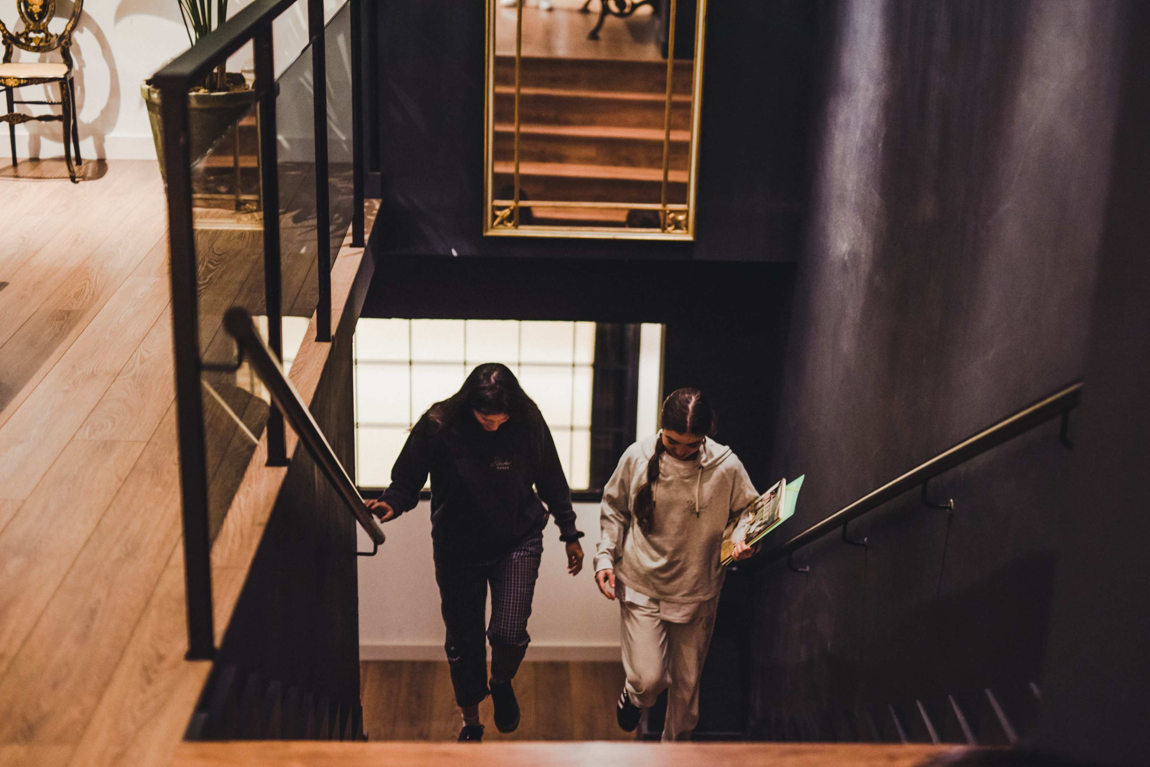 Two women walk down a staircase in a modern interior, with wooden steps and a dark wall.