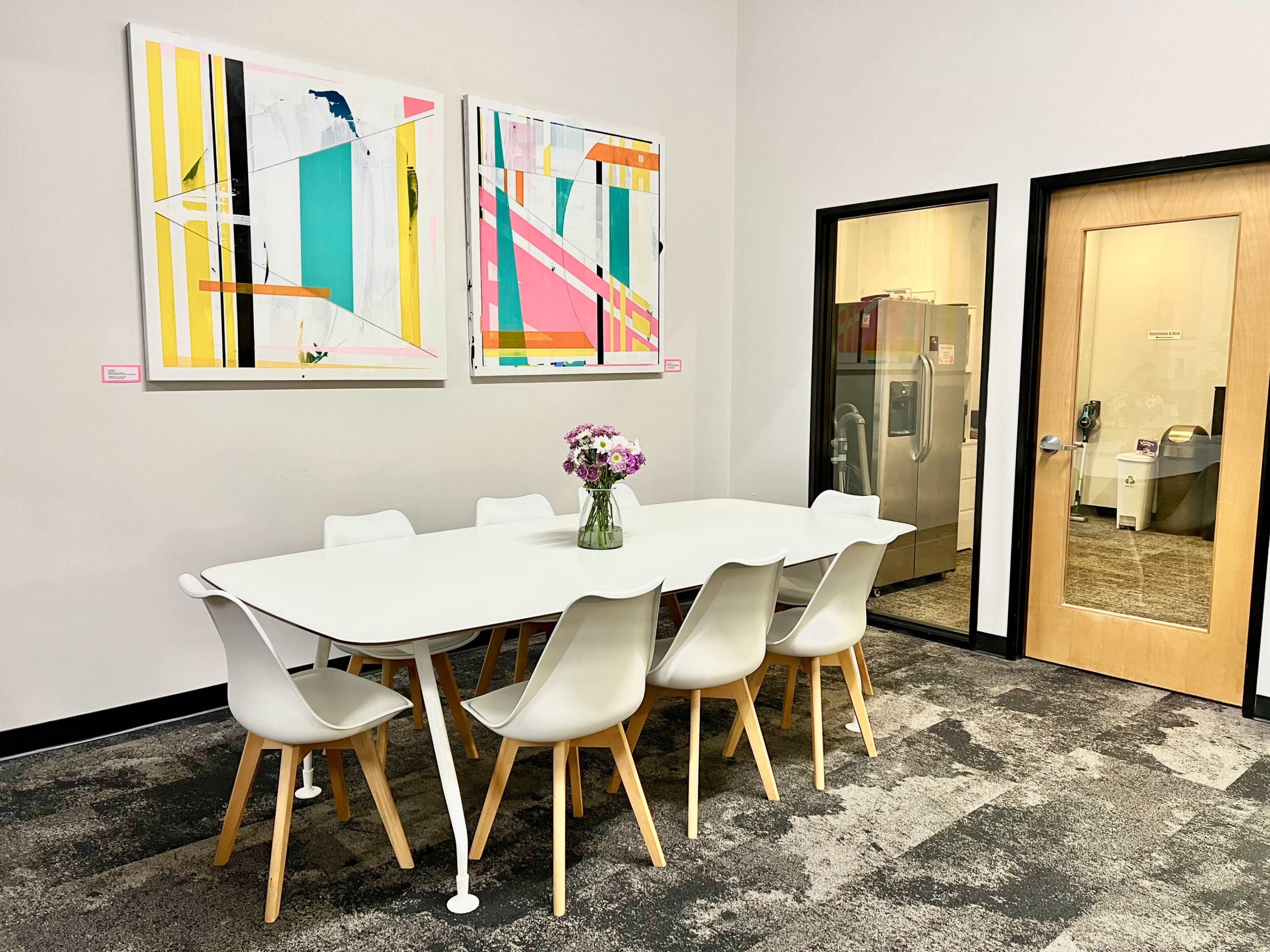 The image shows a modern conference room featuring a white table surrounded by eight white chairs, with two colorful abstract paintings on the wall and a glass door leading to an area with appliances.