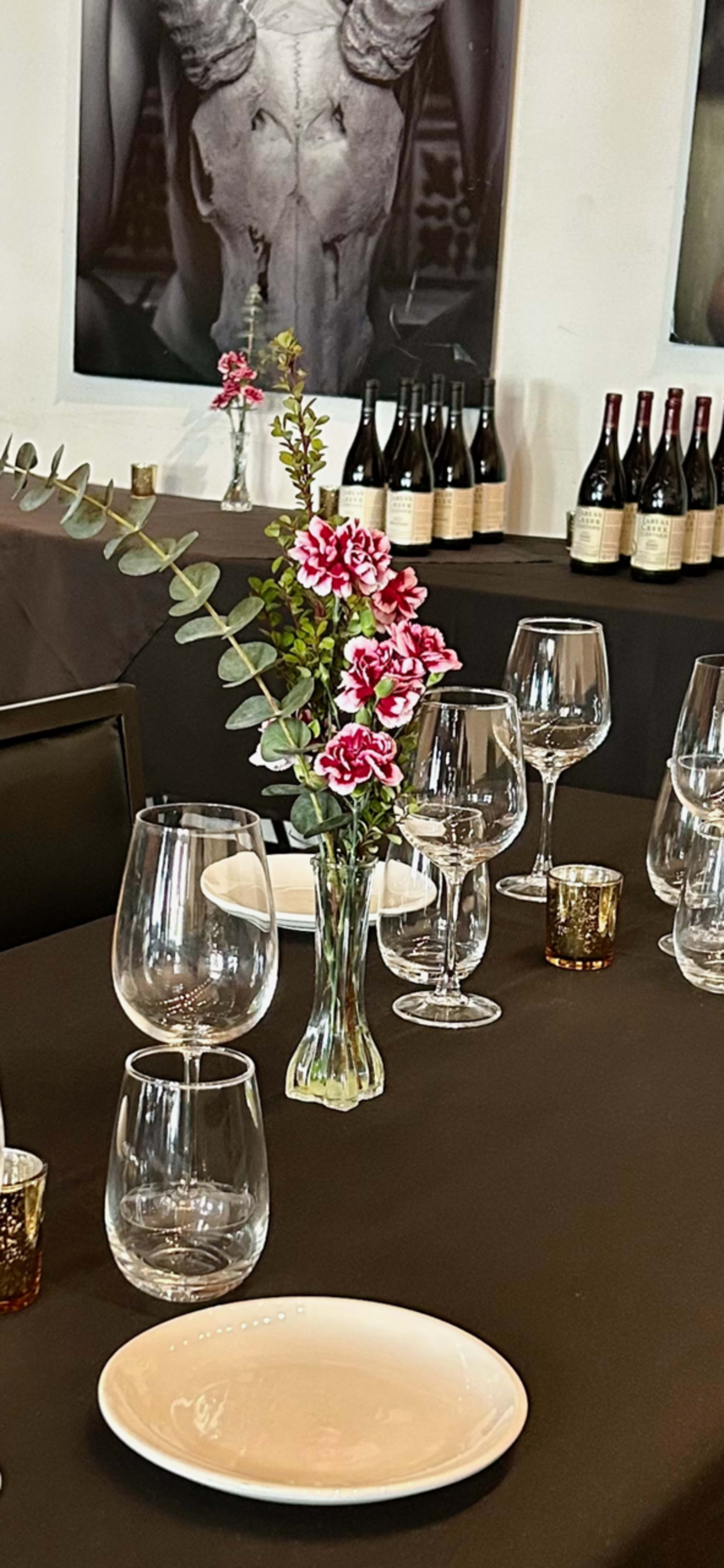 A dining table is set with wine glasses, plates, and a vase of pink flowers, accompanied by bottles of wine in the background.