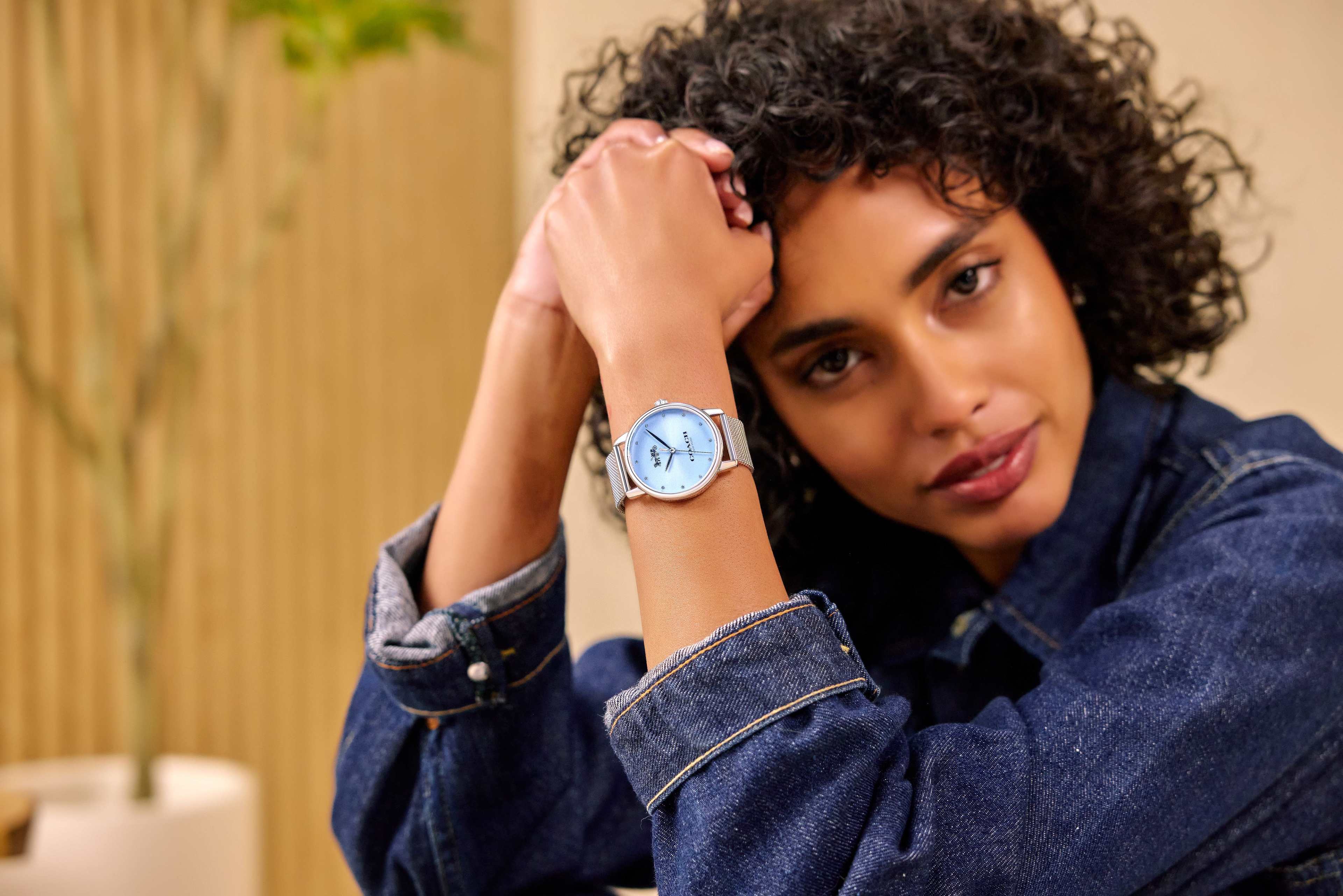 A woman with curly hair is posing while wearing a blue watch and a denim jacket.