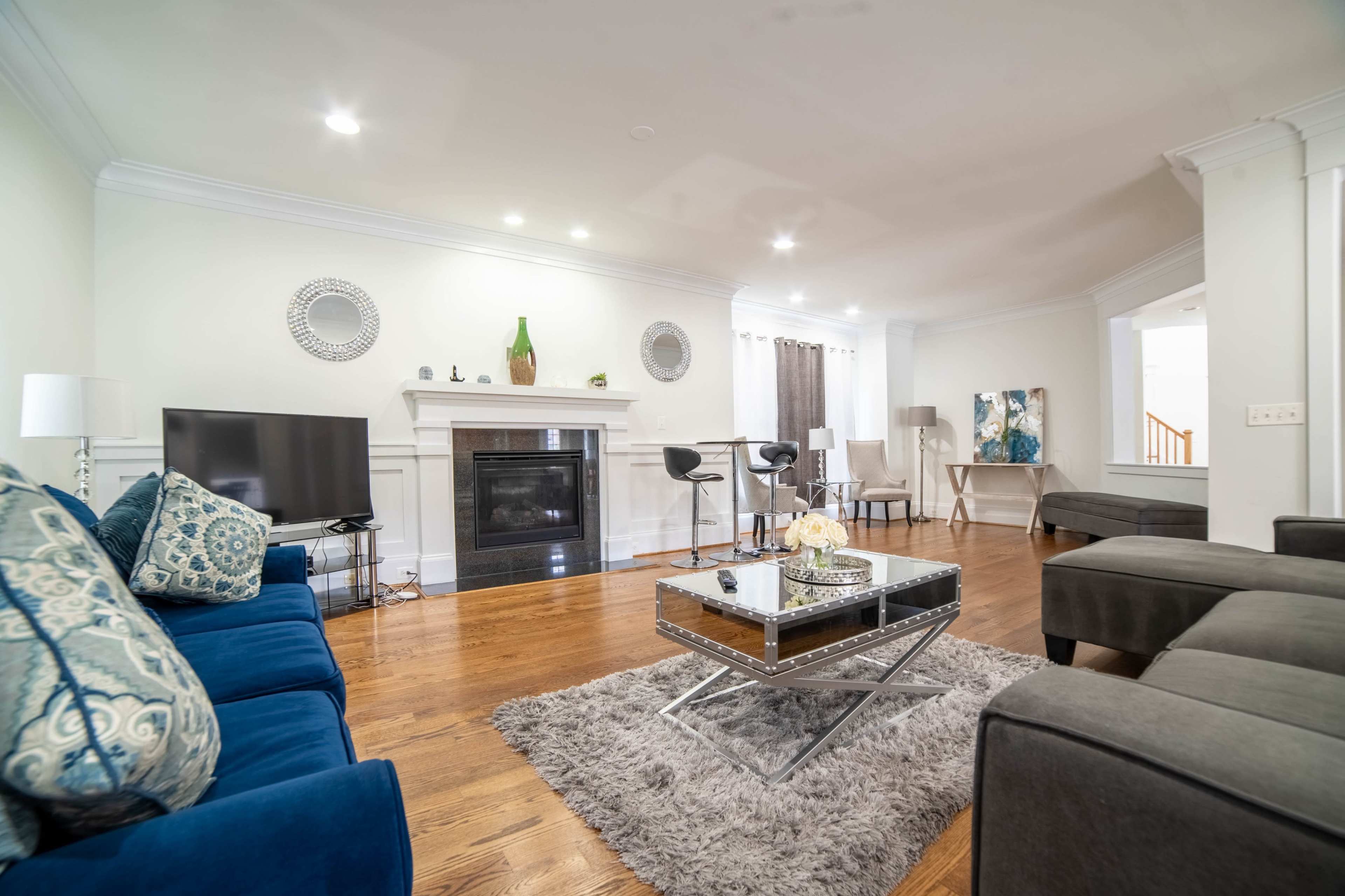 A modern living room features a fireplace, gray area rug, blue sofa, and a glass coffee table, with various seating arrangements and decorative items.