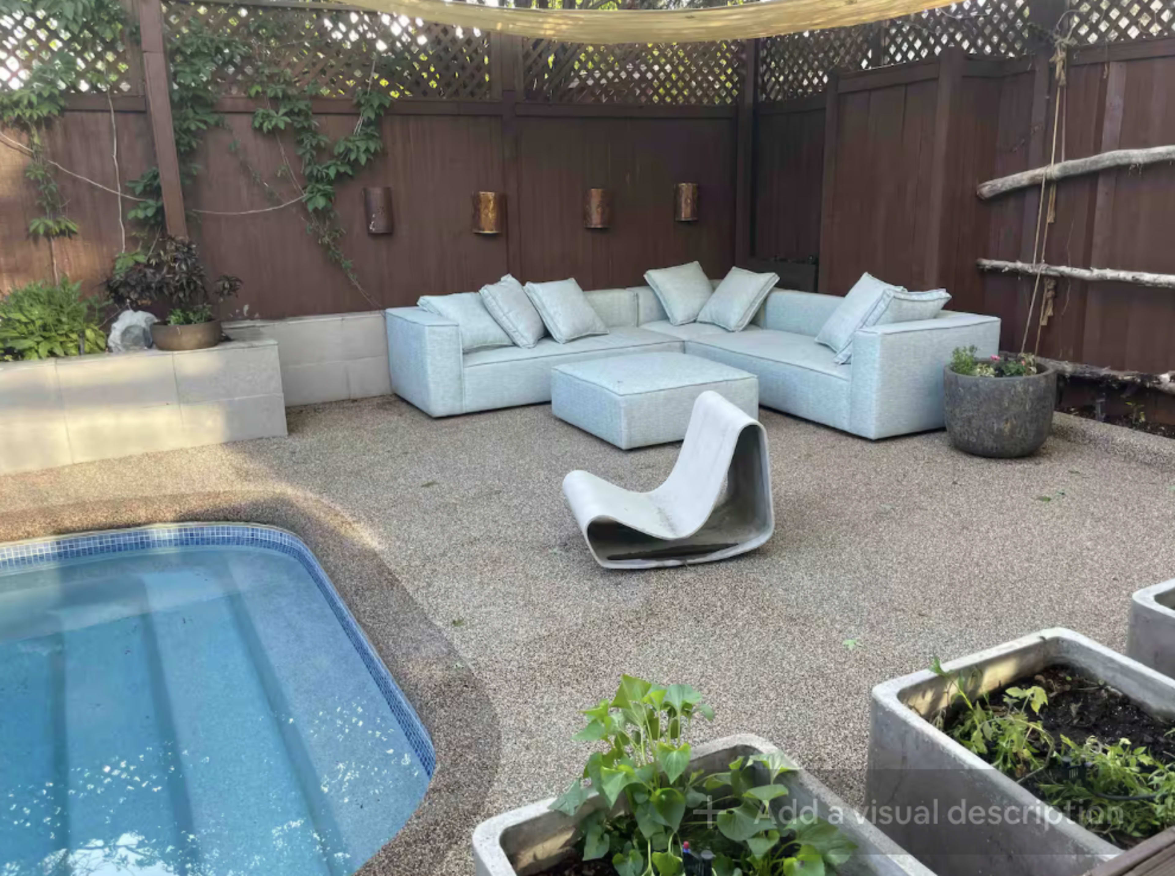 The image shows a backyard patio area featuring a small pool, a modern light-colored sectional sofa with cushions, a minimalist chair, and potted plants arranged around the space.