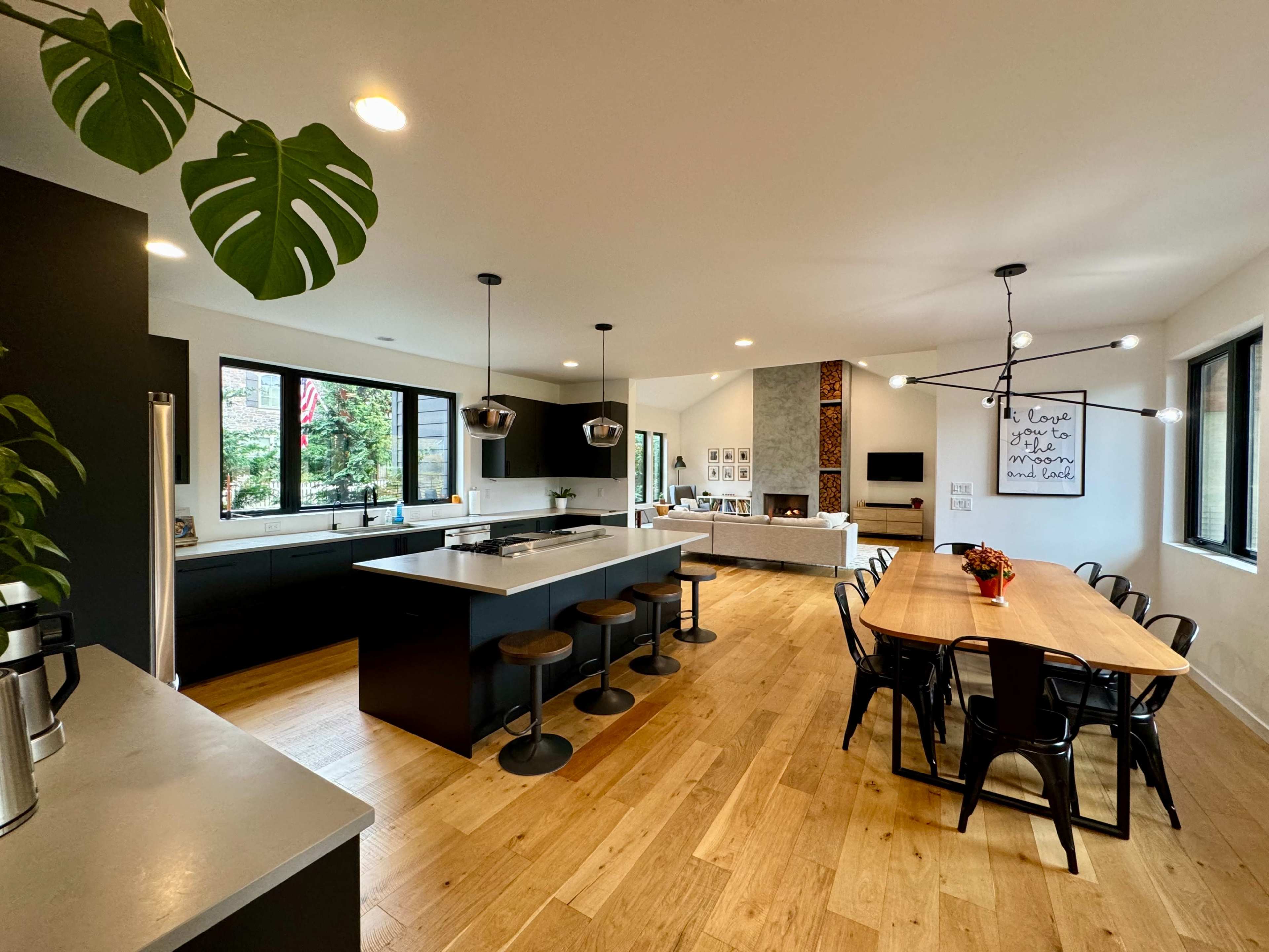 A modern kitchen and dining area featuring a central island with seating, dark cabinetry, a wooden dining table, and large windows allowing natural light.