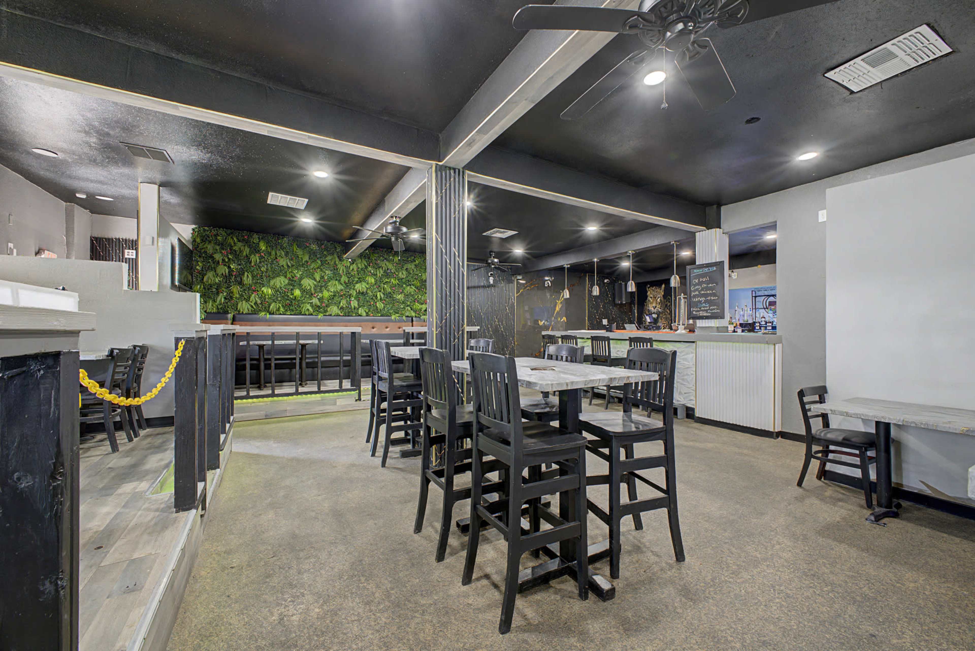 The image shows the interior of a restaurant with wooden tables and black chairs arranged in a spacious layout, featuring a green wall and a bar area in the background.