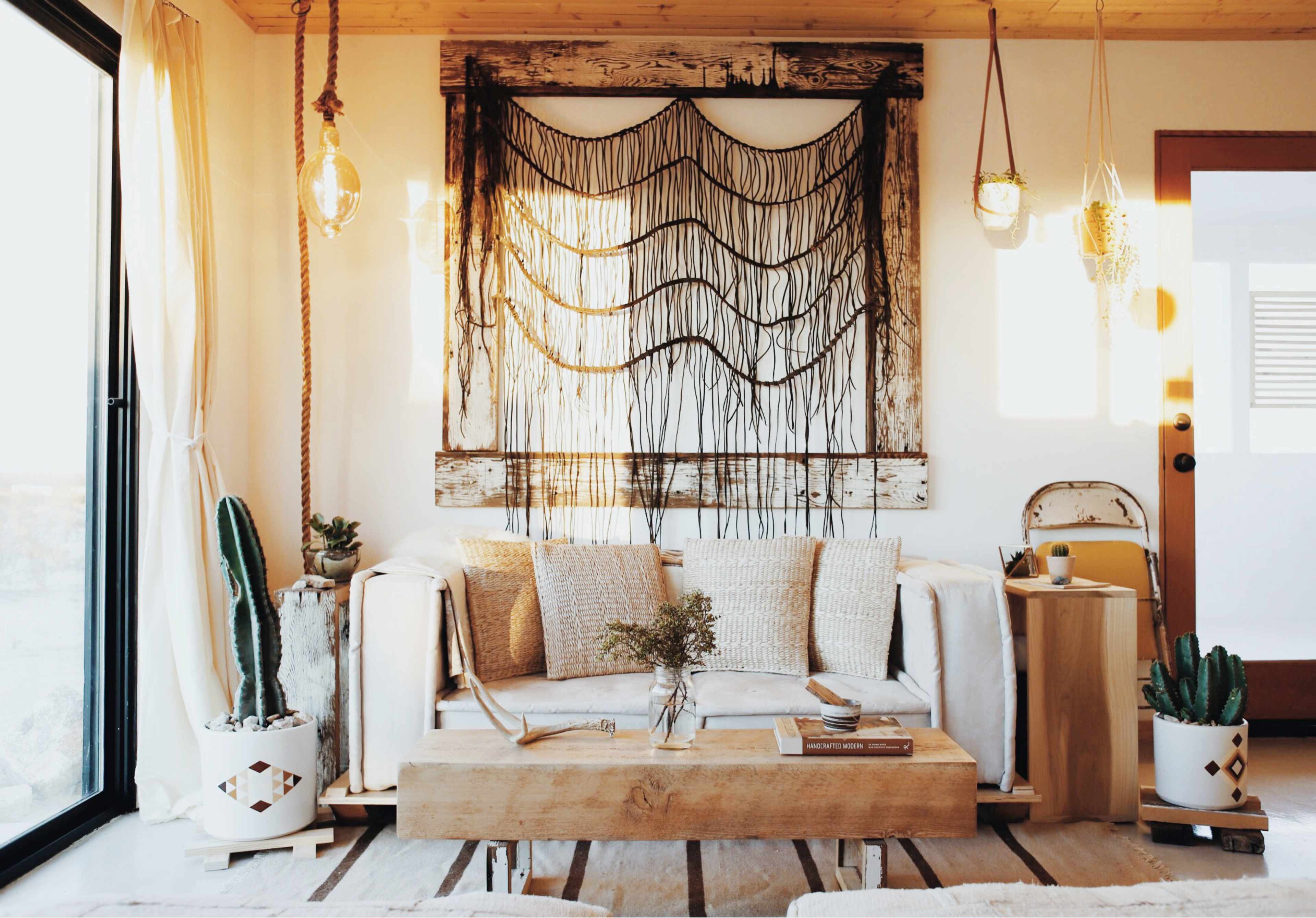 The image shows a living room with a white couch, a wooden coffee table, and a wall adorned with a macramé art piece, illuminated by natural light from large windows.