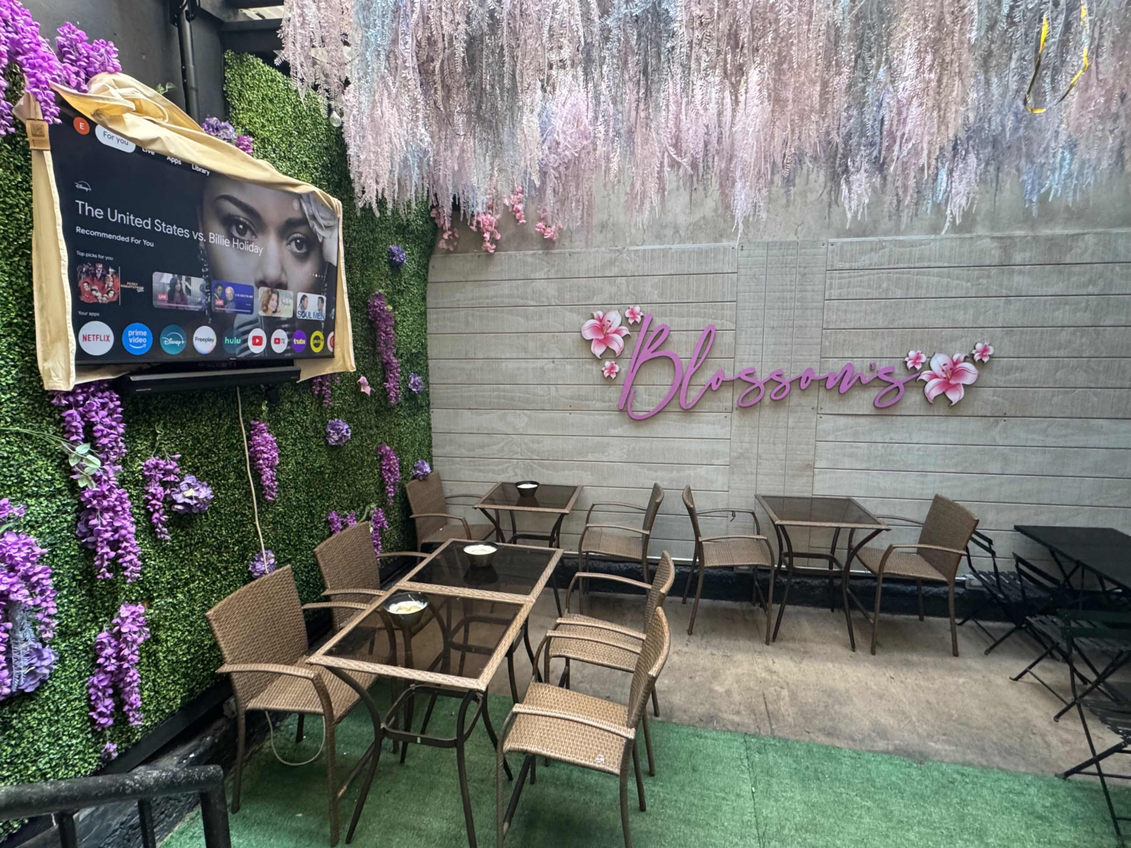 A small outdoor dining area features several tables with brown wicker chairs, surrounded by artificial greenery and decorative flowers, and includes a wall-mounted screen displaying content.