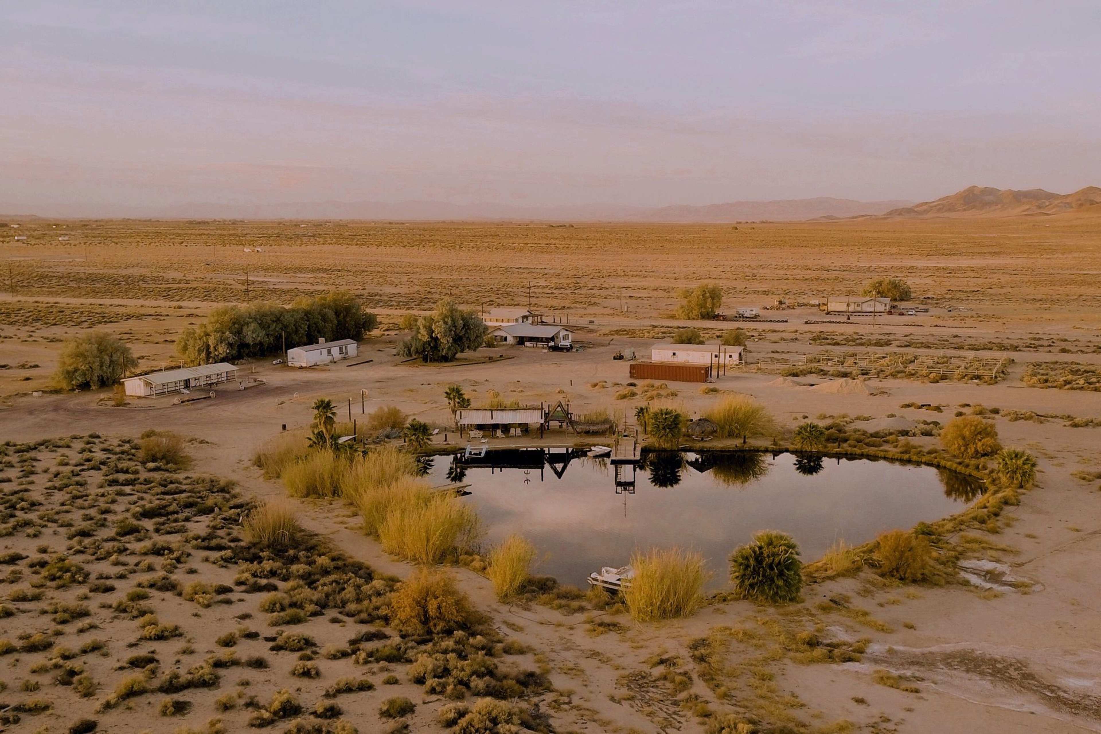 A remote desert landscape with several trailers and a pond surrounded by sparse vegetation.
