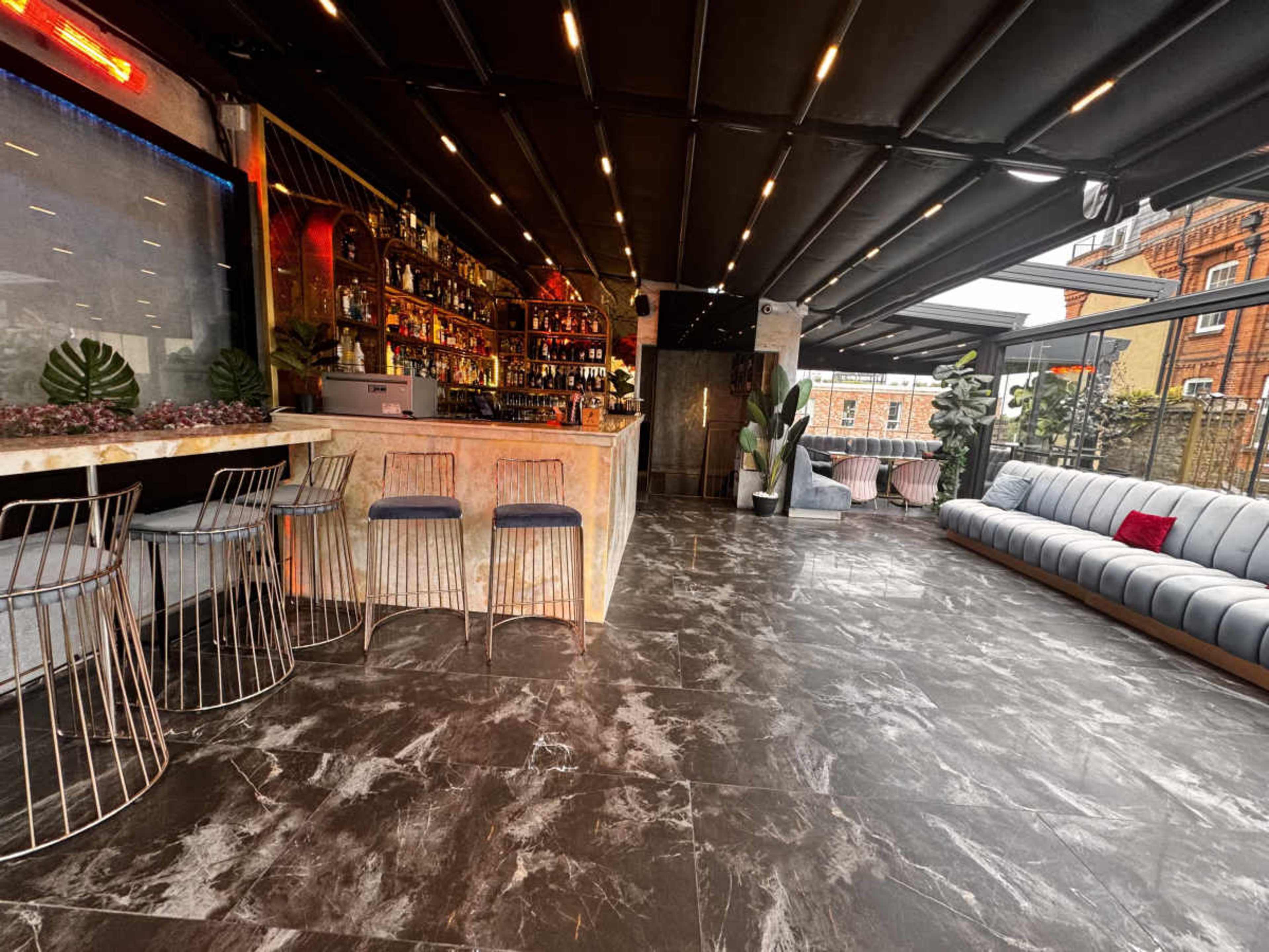 The image shows a modern rooftop bar with a well-stocked bar counter, high stools, and a spacious seating area featuring a long sofa.