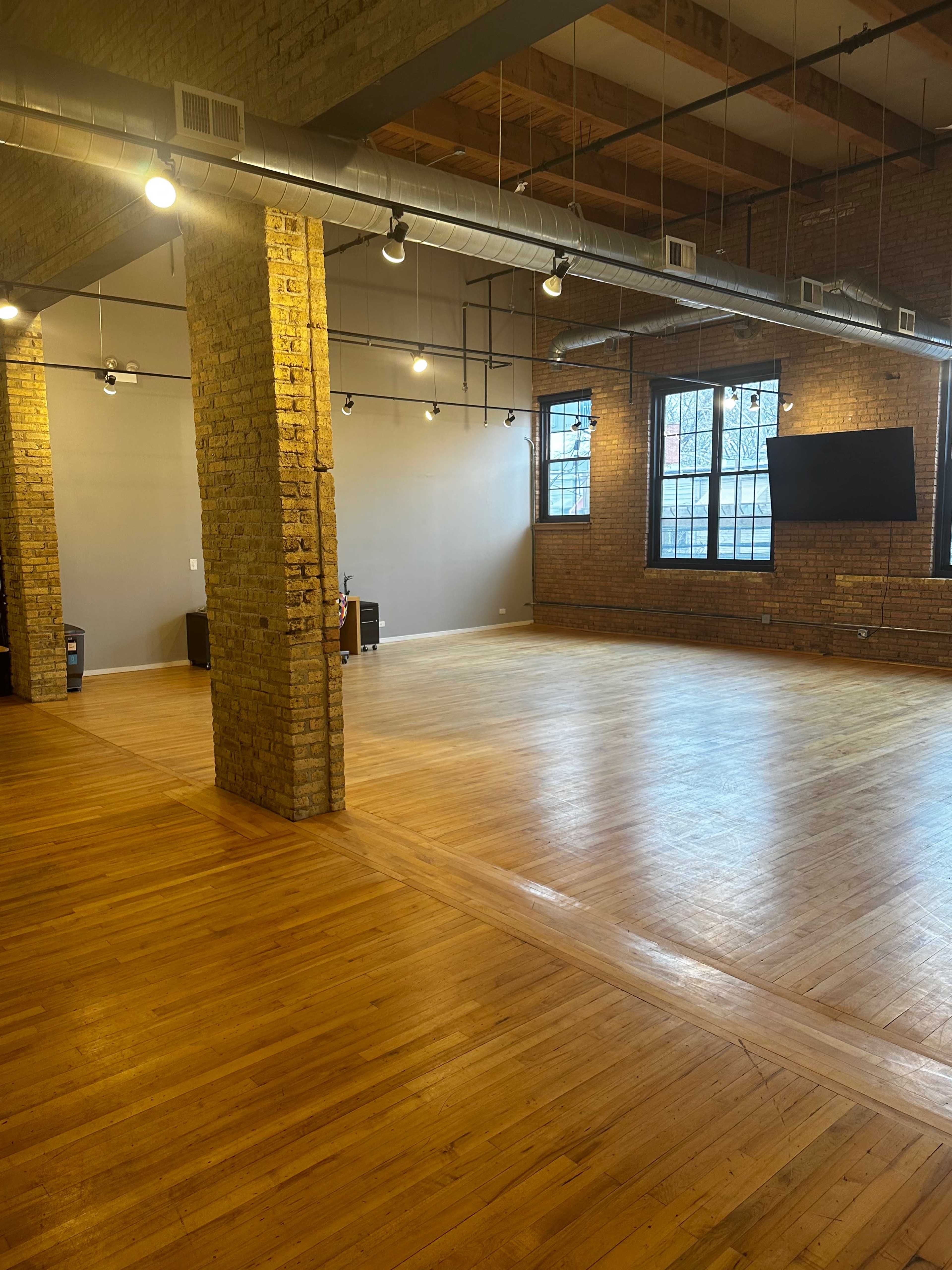 An empty interior space with wooden floors, exposed brick walls, and large windows, featuring track lighting and a mounted screen.