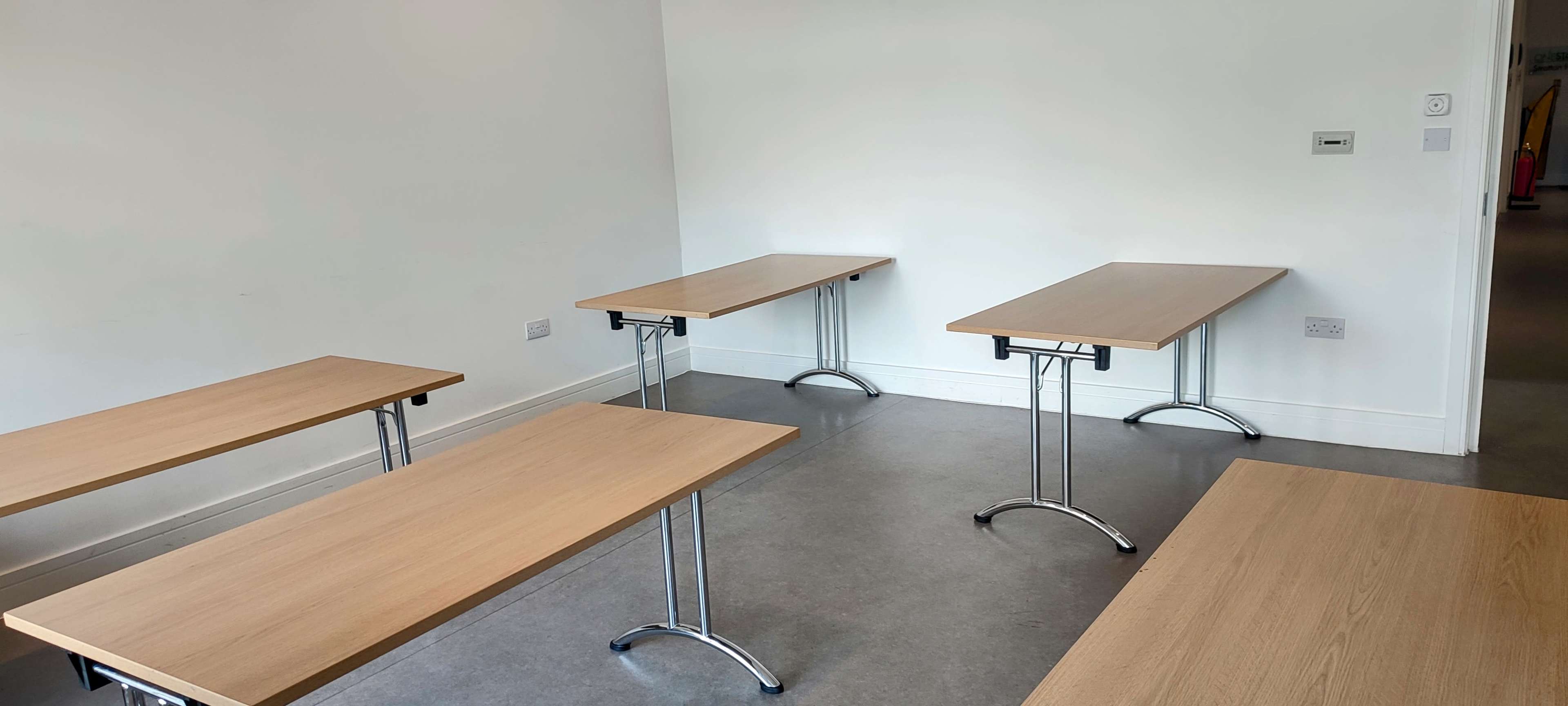 A room with several wooden tables arranged in a sparse layout against a plain white wall.