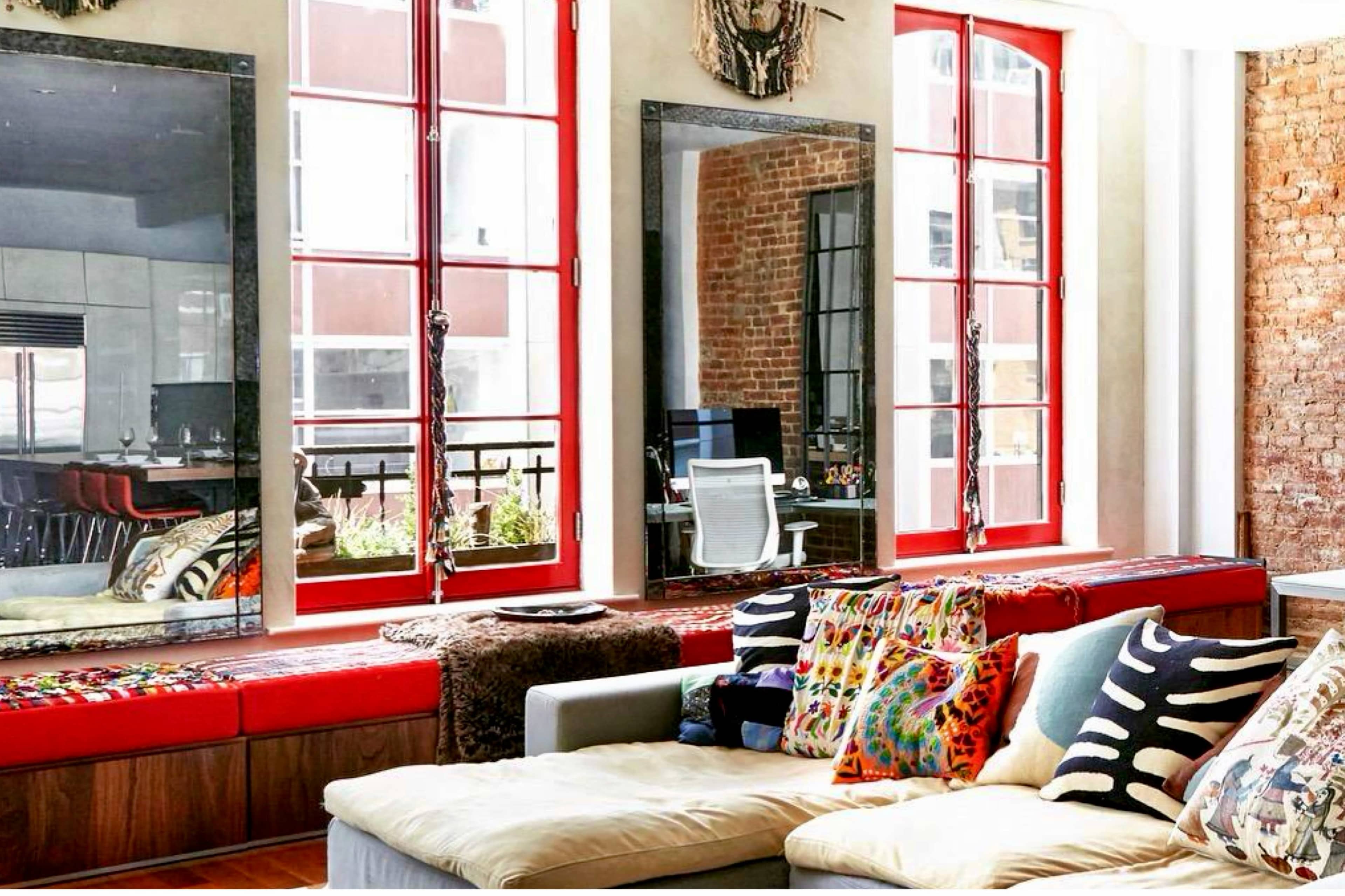 The image shows a bright living space with large red-framed windows, a beige sectional couch adorned with colorful pillows, and a workspace visible in the background.