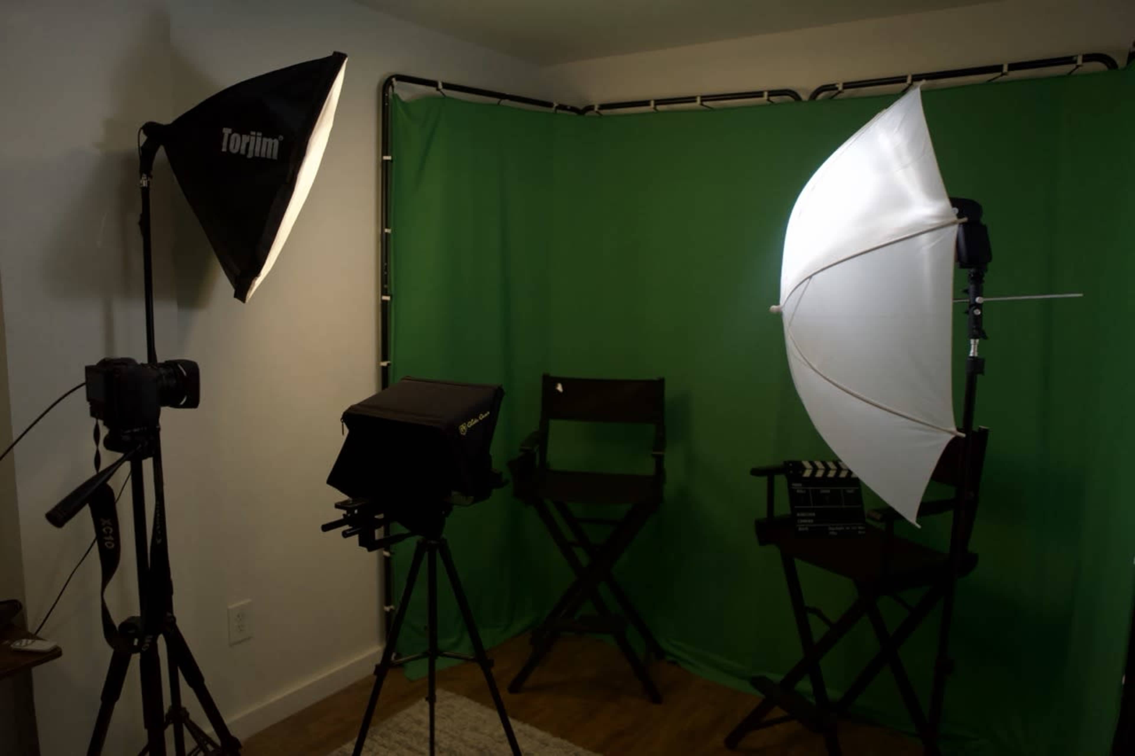 The image shows a small studio setup featuring a green backdrop, two lighting equipment stands, a camera on a tripod, and two director's chairs.