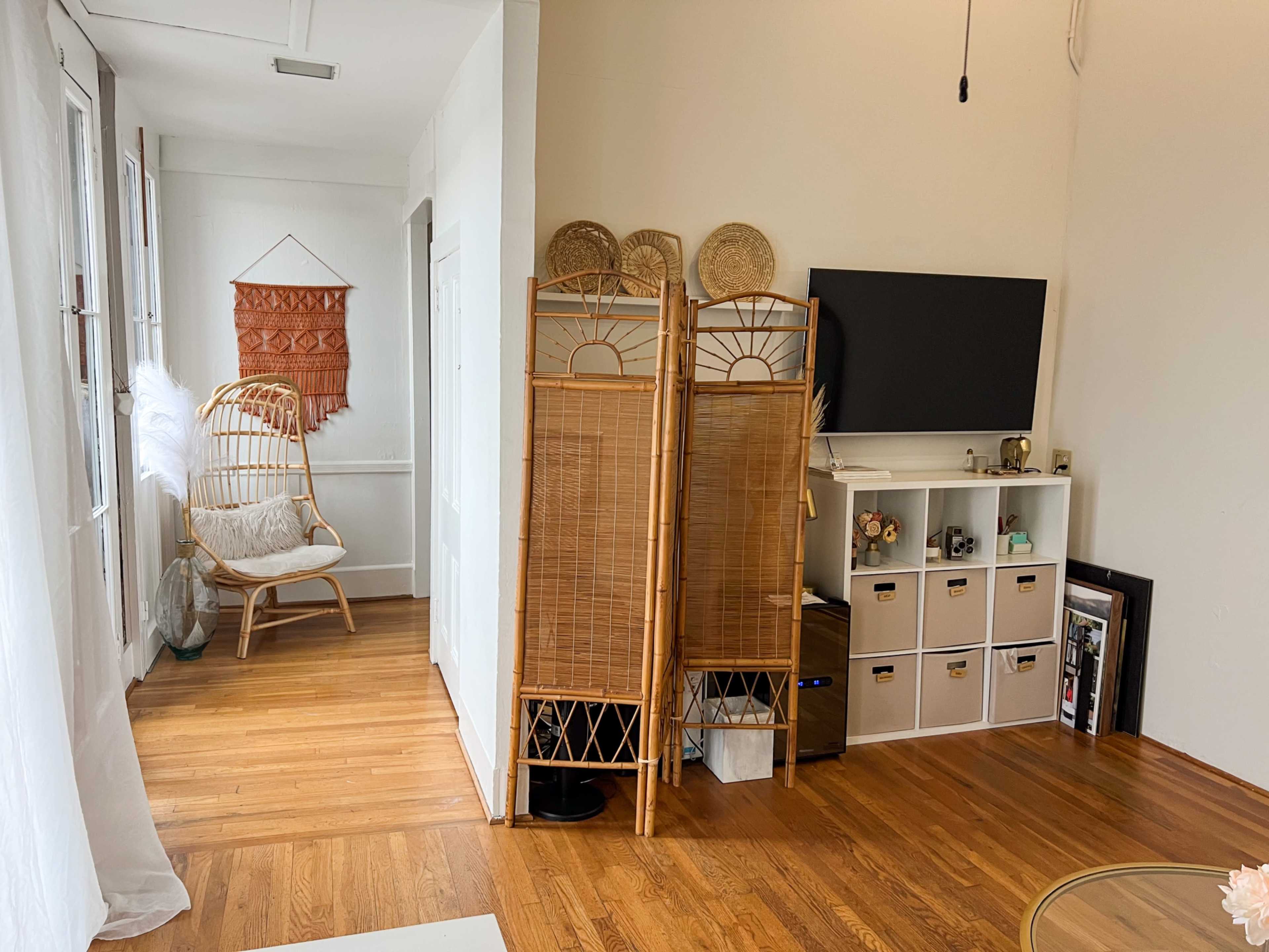 The image depicts a bright room featuring wooden floors, a rattan chair, two folding screens, a television, and a shelving unit with various items.
