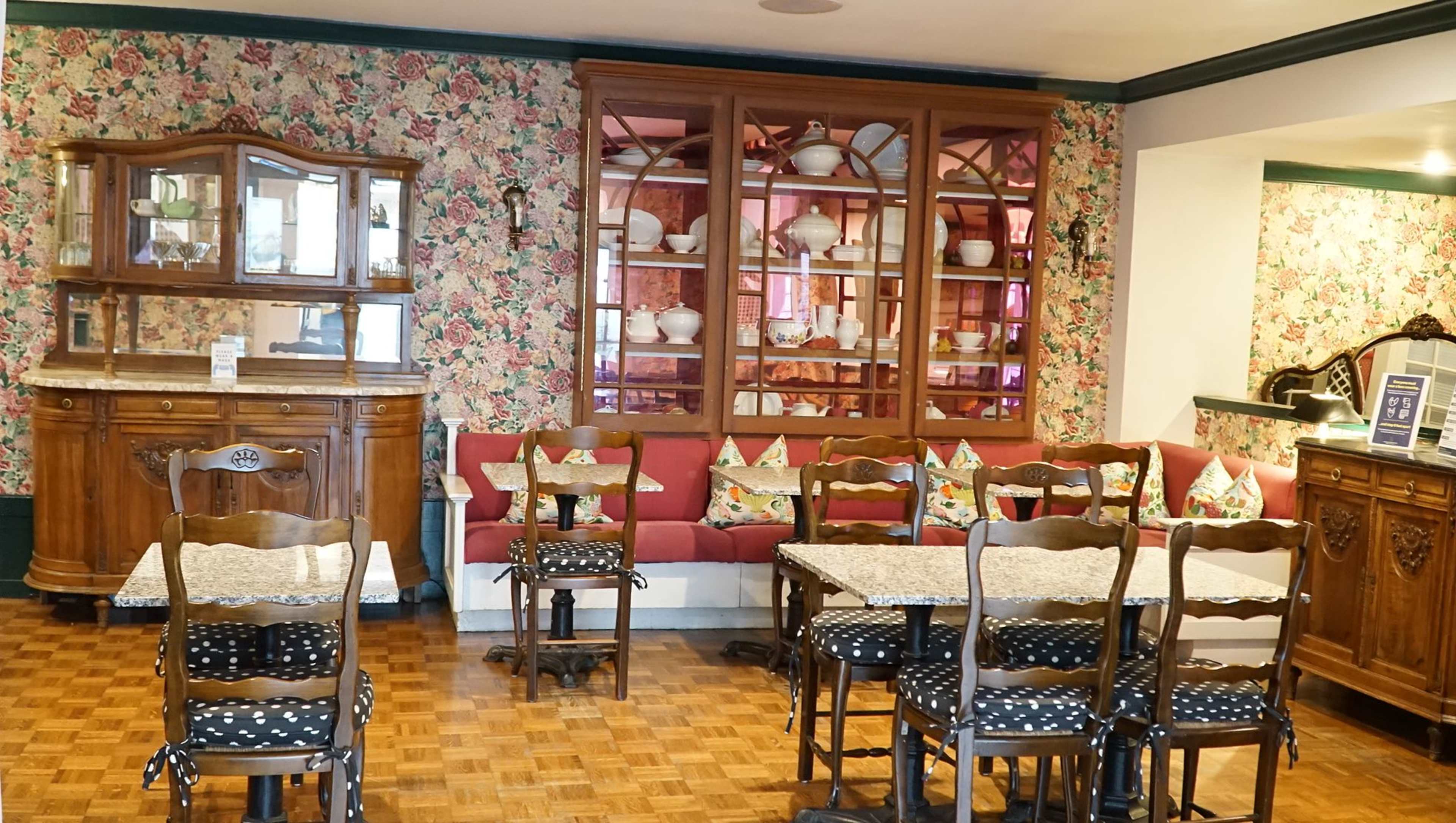 The image shows a cozy café interior featuring floral wallpaper, wooden furniture, and a display cabinet filled with dishware.