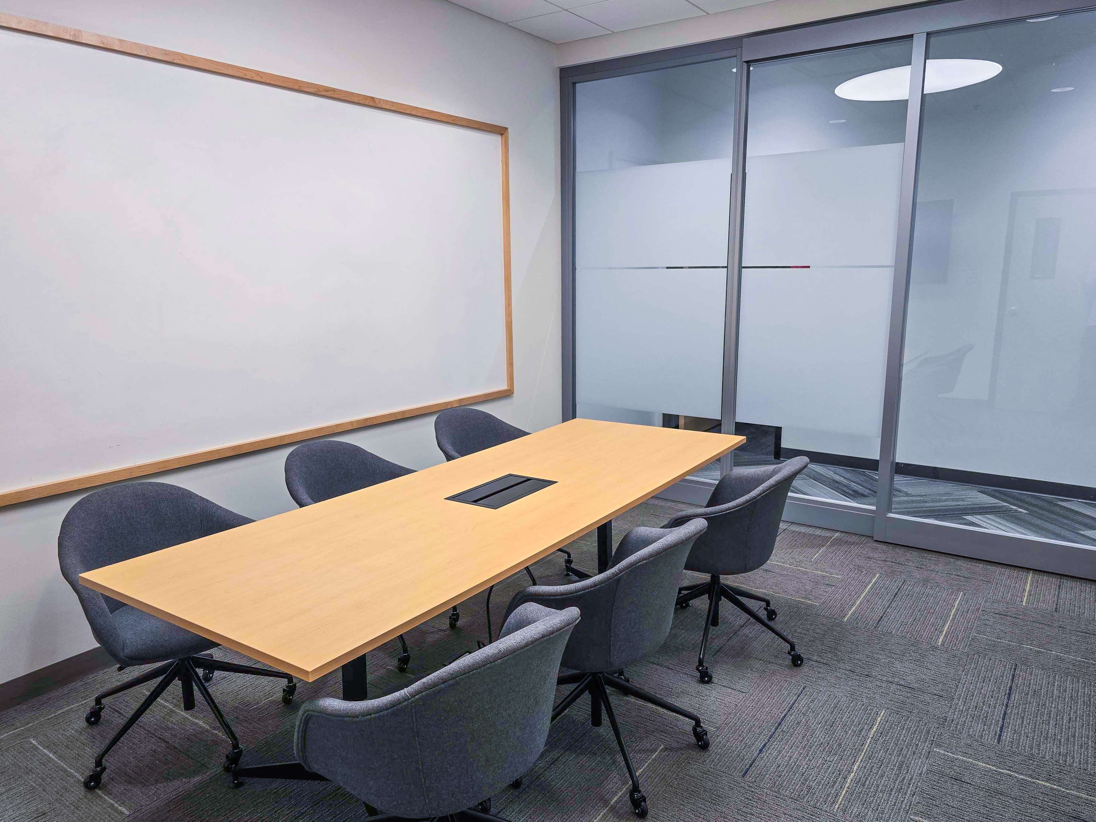 The image shows a modern conference room equipped with a large wooden table and six rolling chairs, alongside a wall-mounted whiteboard and glass doors.