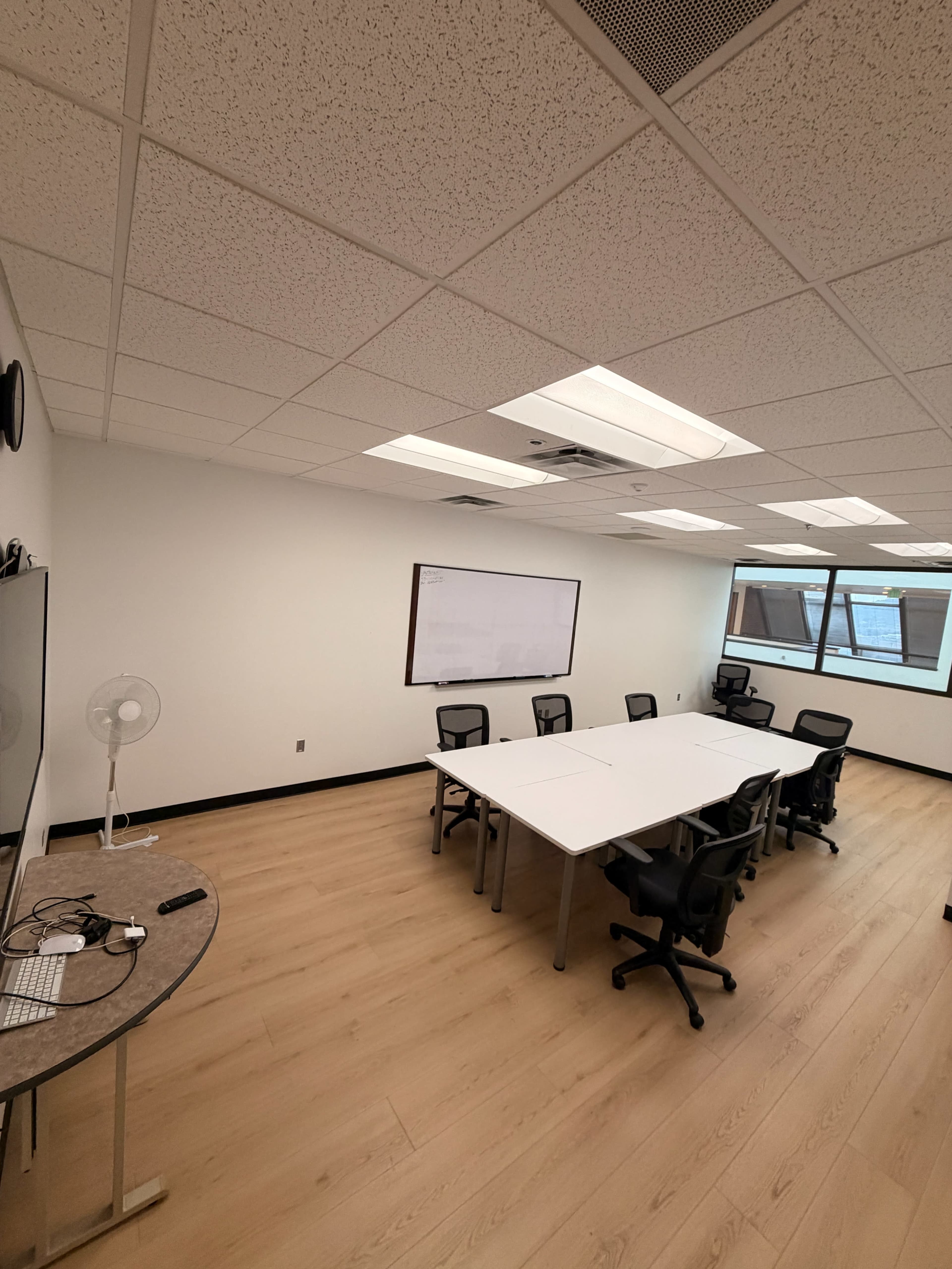 The image shows a modern conference room with a long rectangular table surrounded by black chairs, a whiteboard on the wall, and a fan in the corner.