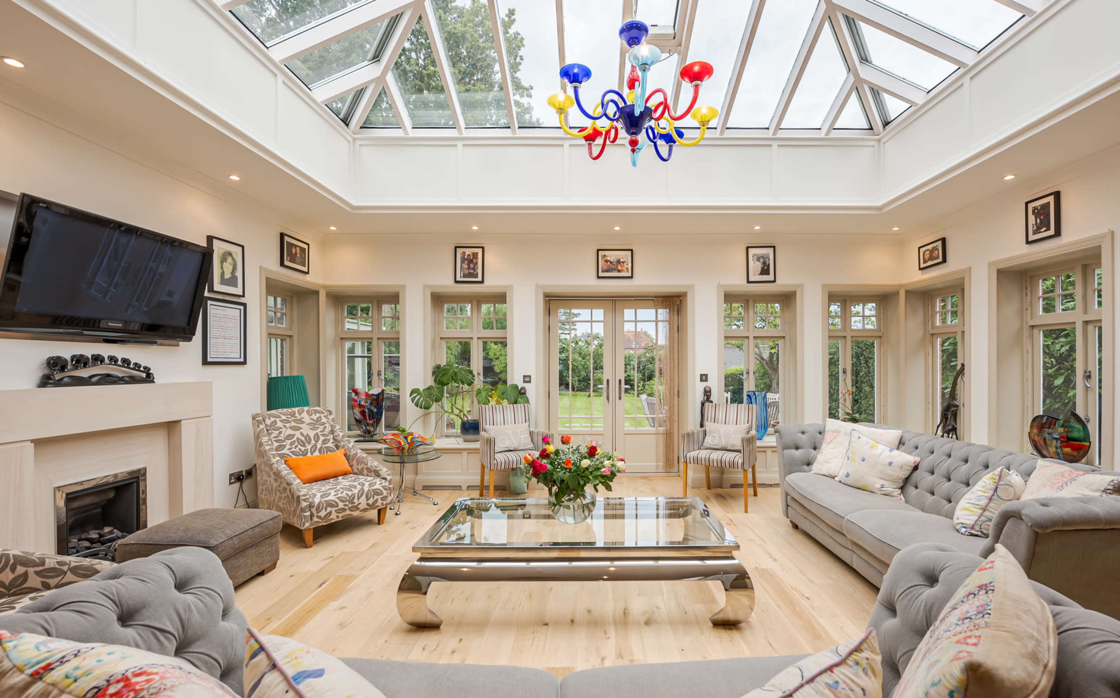 A bright conservatory with a glass ceiling, featuring a spacious seating area with a coffee table and a view of a garden outside.