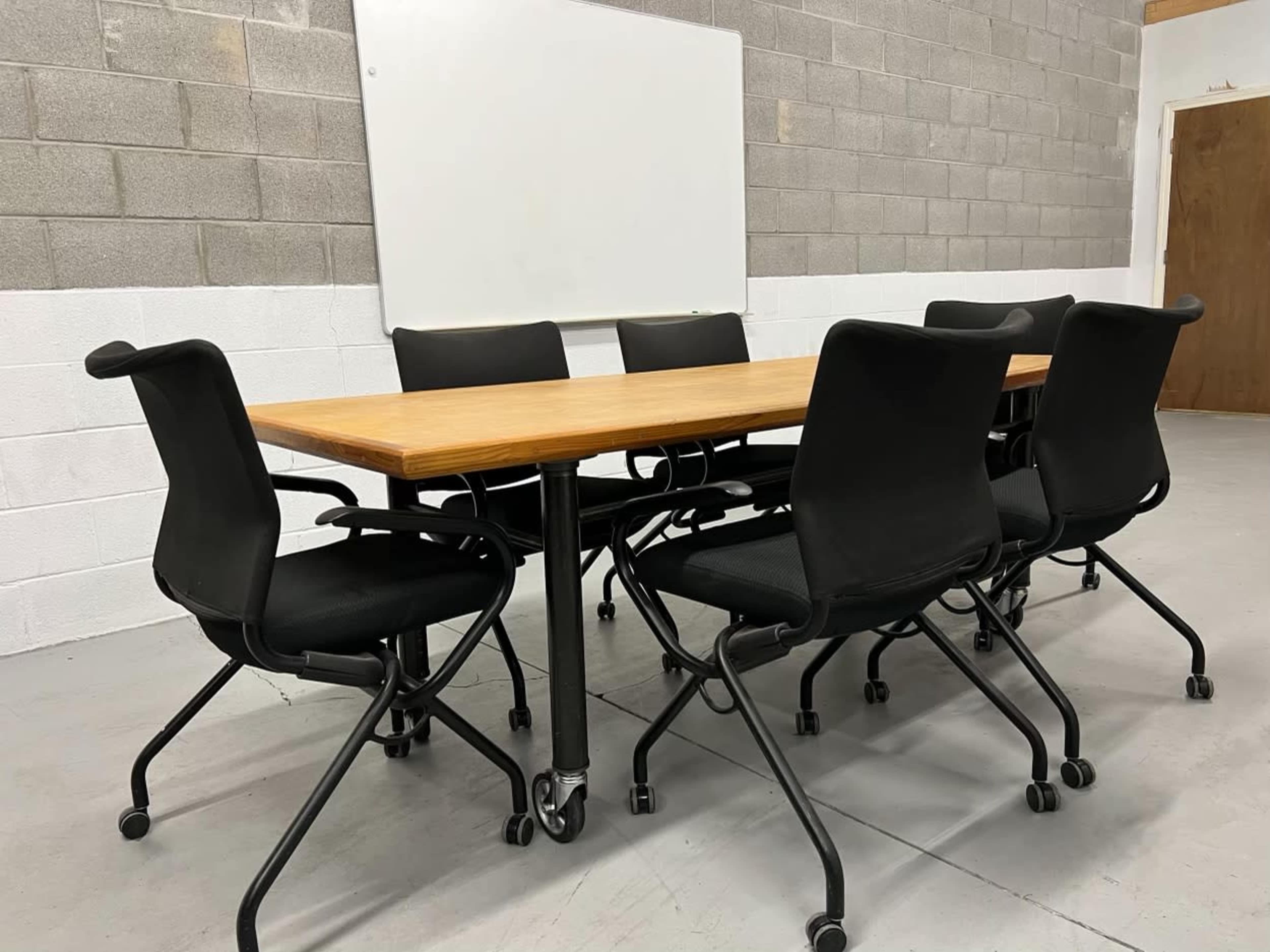A wooden conference table surrounded by six black rolling chairs in a room with gray block walls.