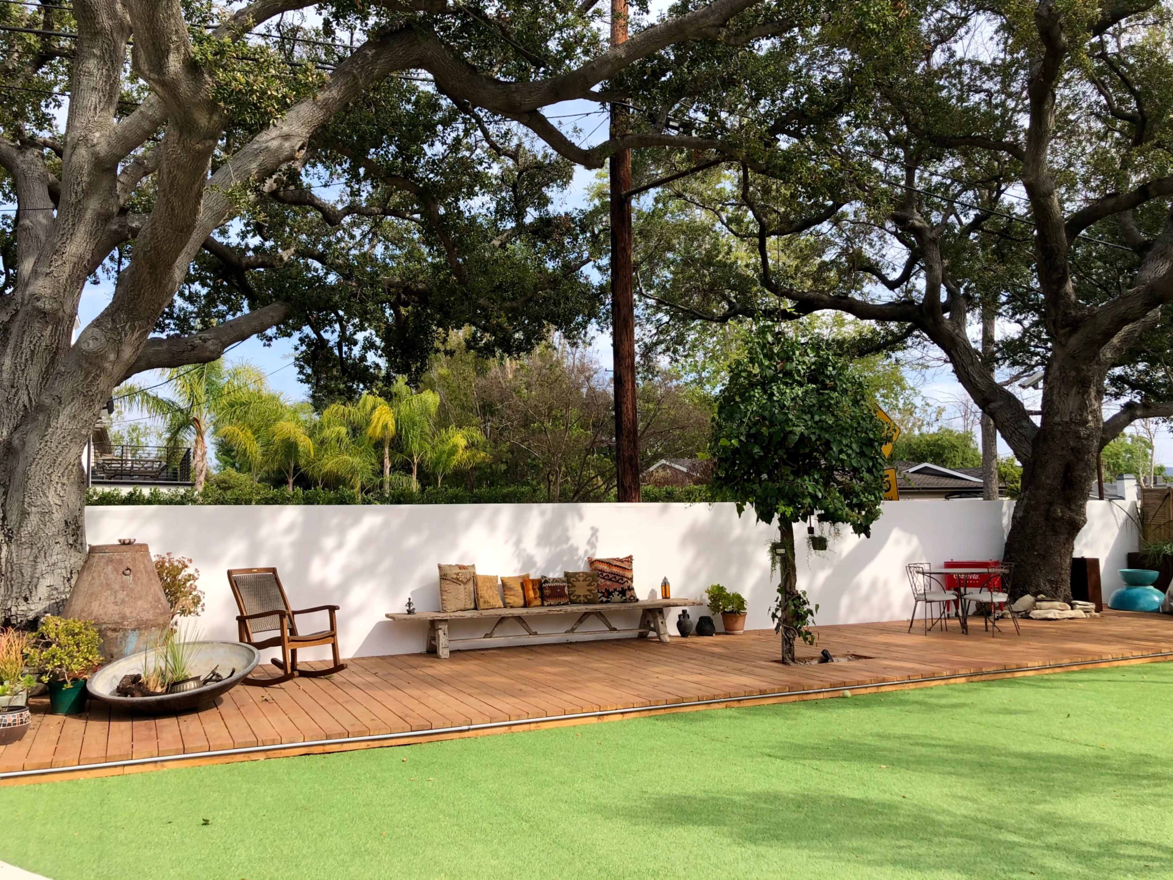 A wooden deck with a bench, a small red table and chairs, and a green lawn bordered by trees and a white wall.