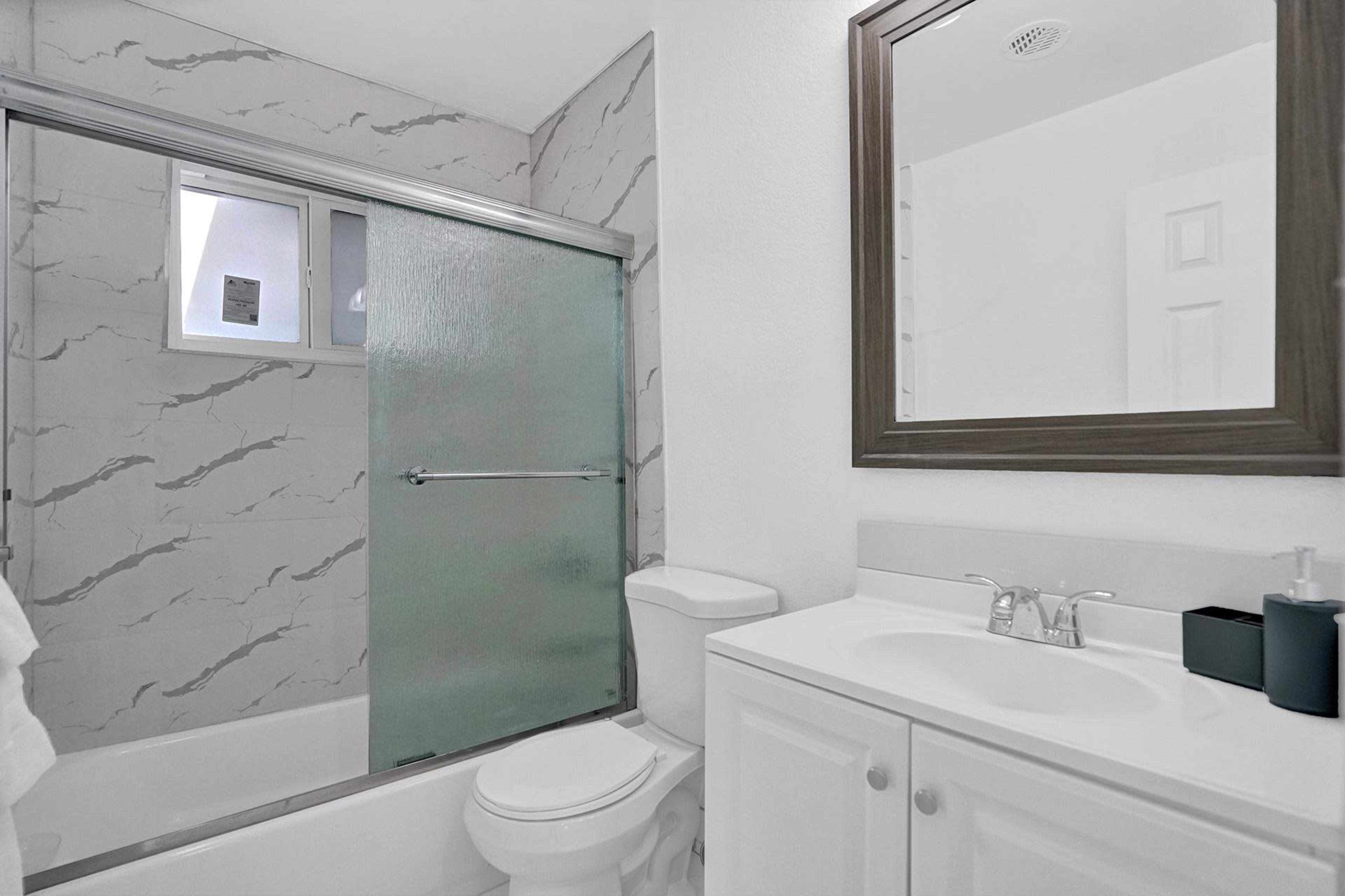 A bathroom with a glass shower, a toilet, and a sink with a mirror above it, featuring light-colored walls and a marble-patterned backsplash.
