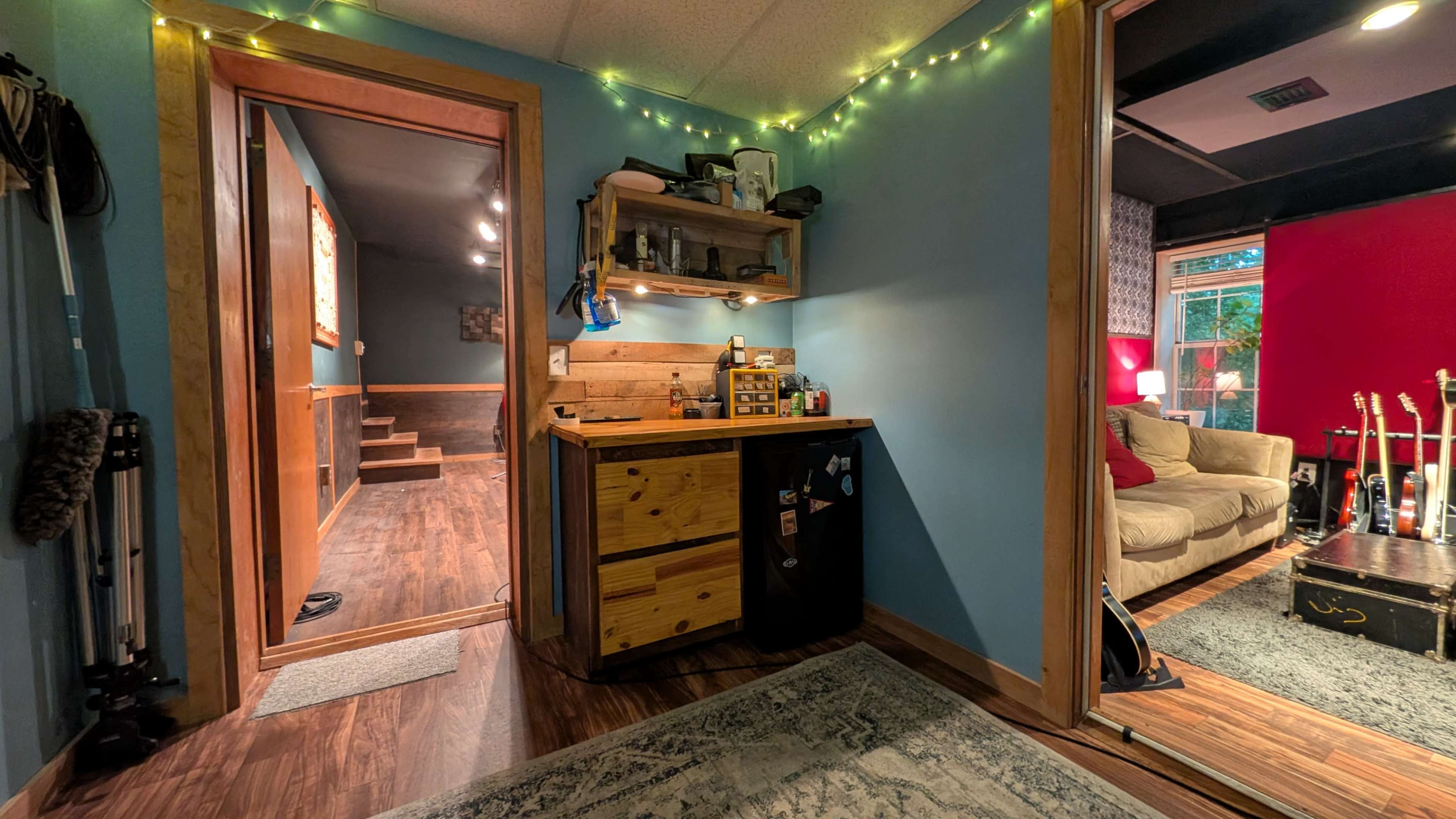 The image shows a cozy interior space featuring a wooden cabinet with a small fridge, a decorated wall with string lights, and an entrance leading to a staircase and another room.