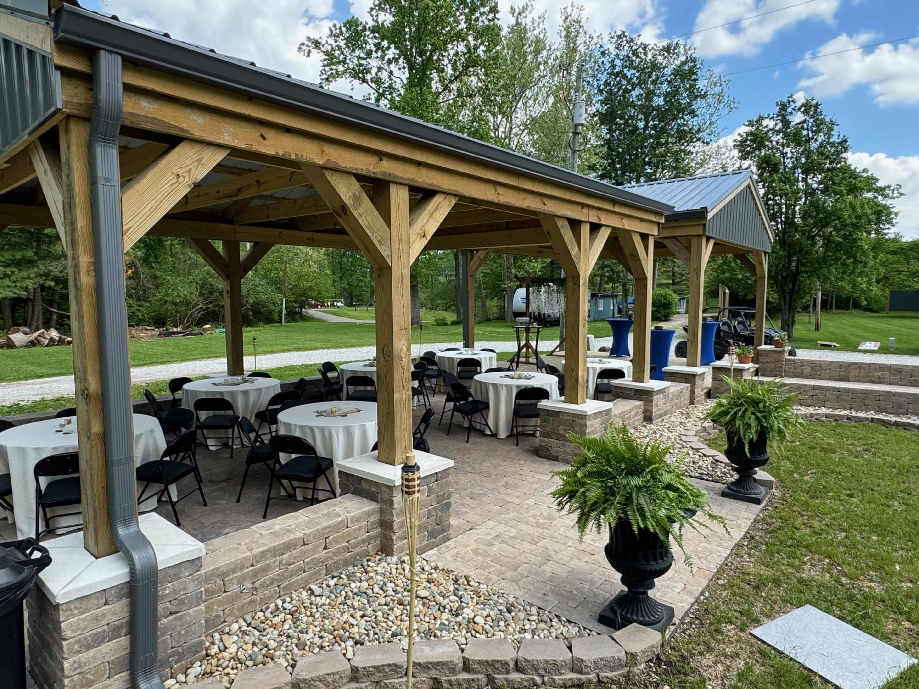 A spacious outdoor pavilion with tables and chairs is set up on a stone patio, surrounded by greenery.