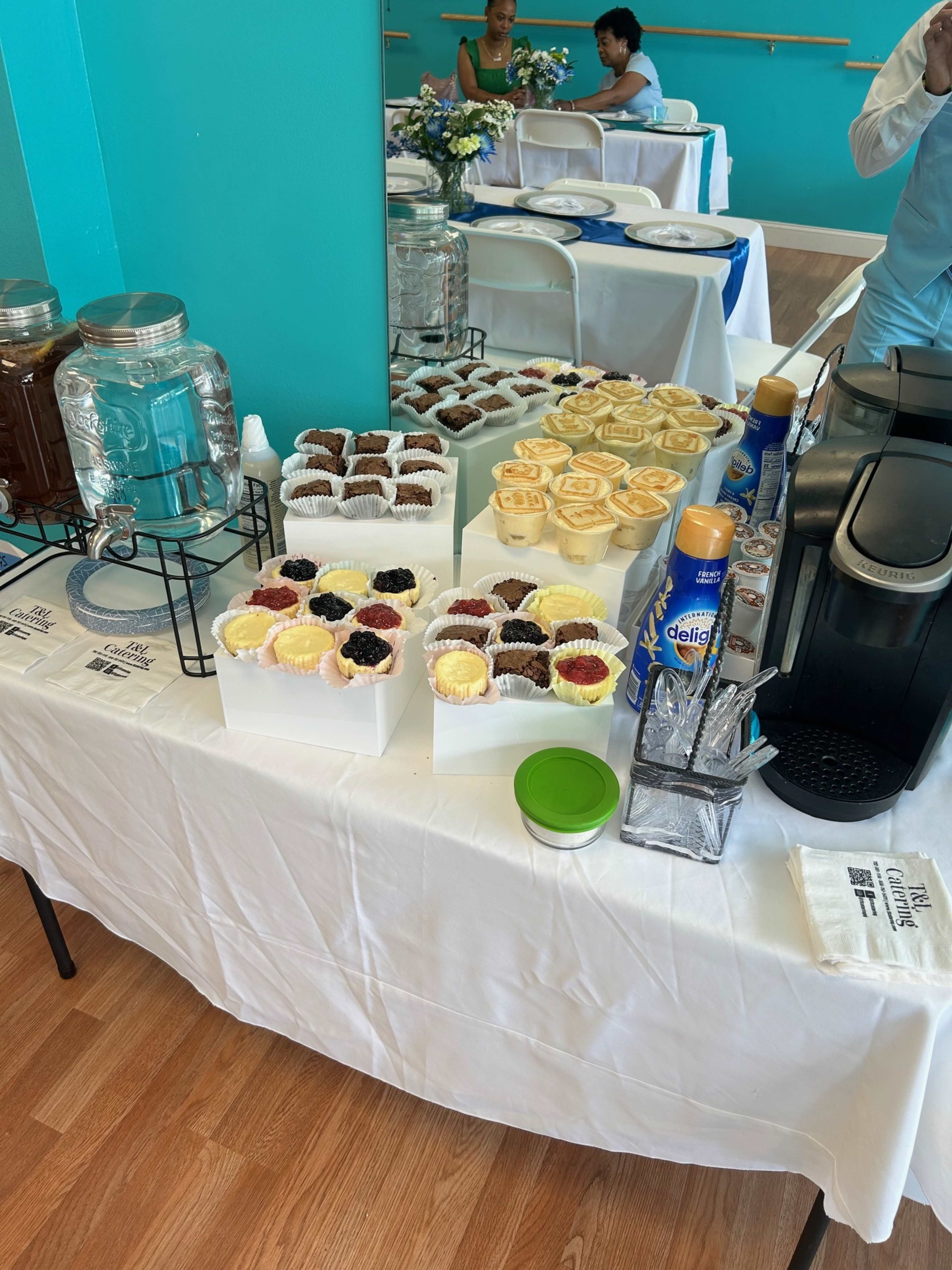 The table displays an assortment of desserts, including cupcakes and small pastries, alongside a beverage station in a brightly colored room.