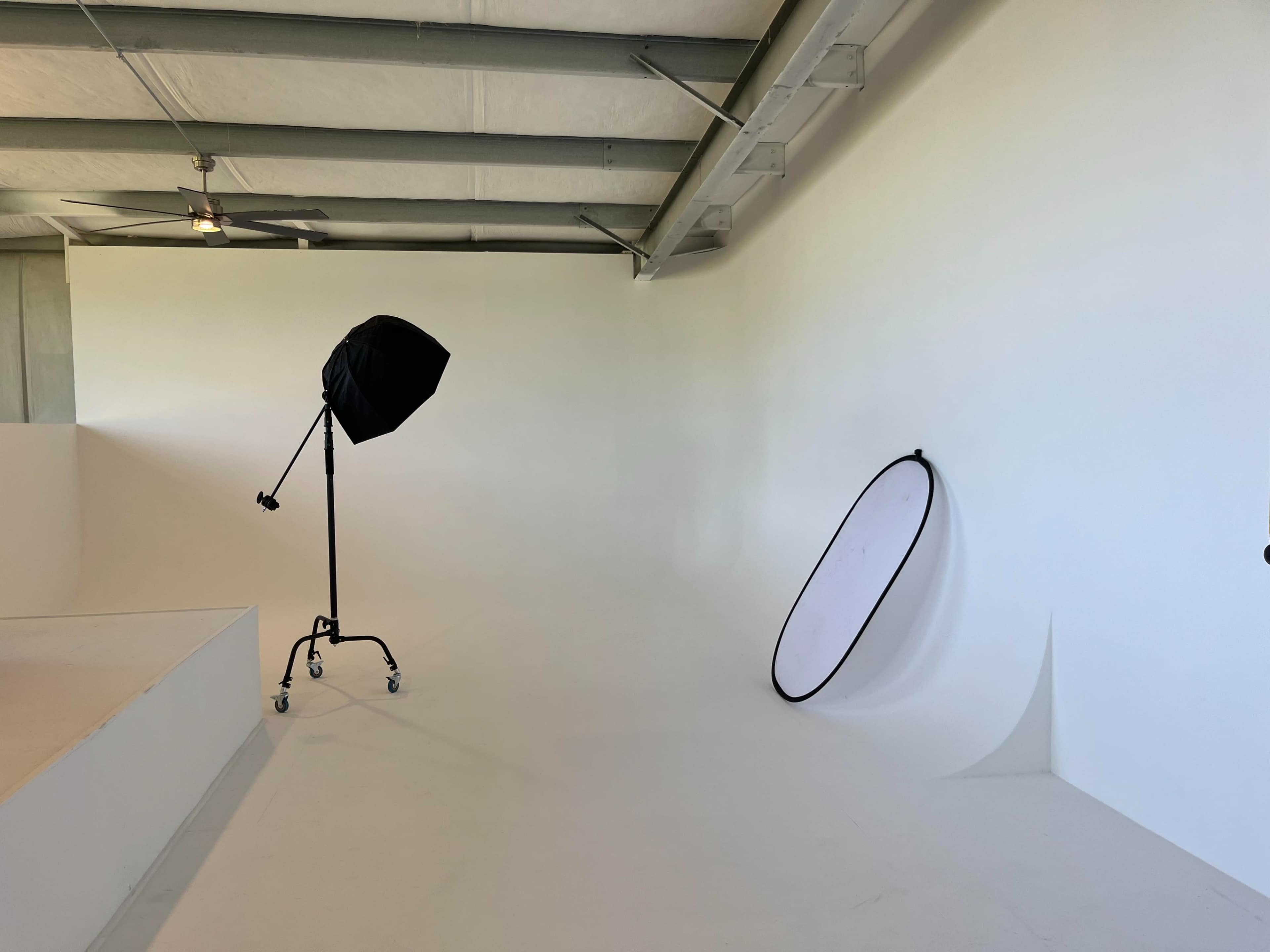 The scene shows a minimalistic photo studio with a softbox light and a large reflector leaning against a white wall.