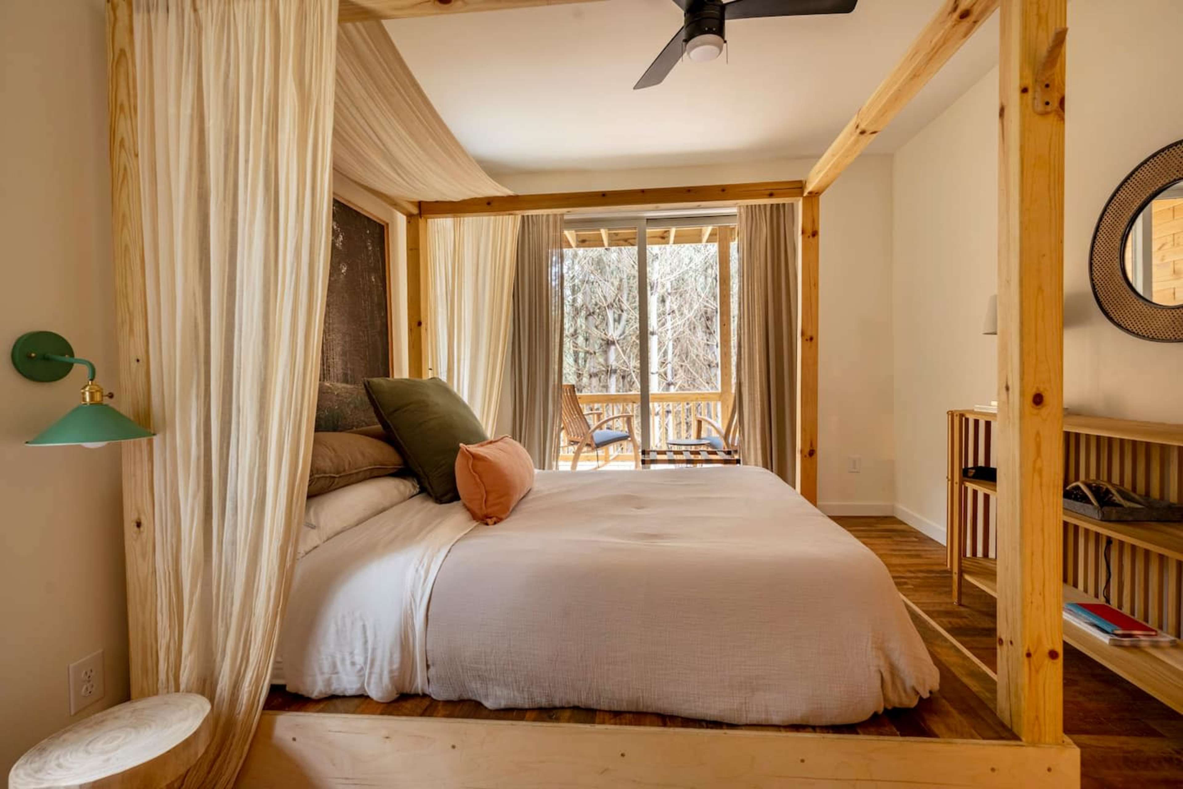 A wooden framed bed with a light-colored bedspread, topped with pillows, in a well-lit room featuring large windows and a balcony view.