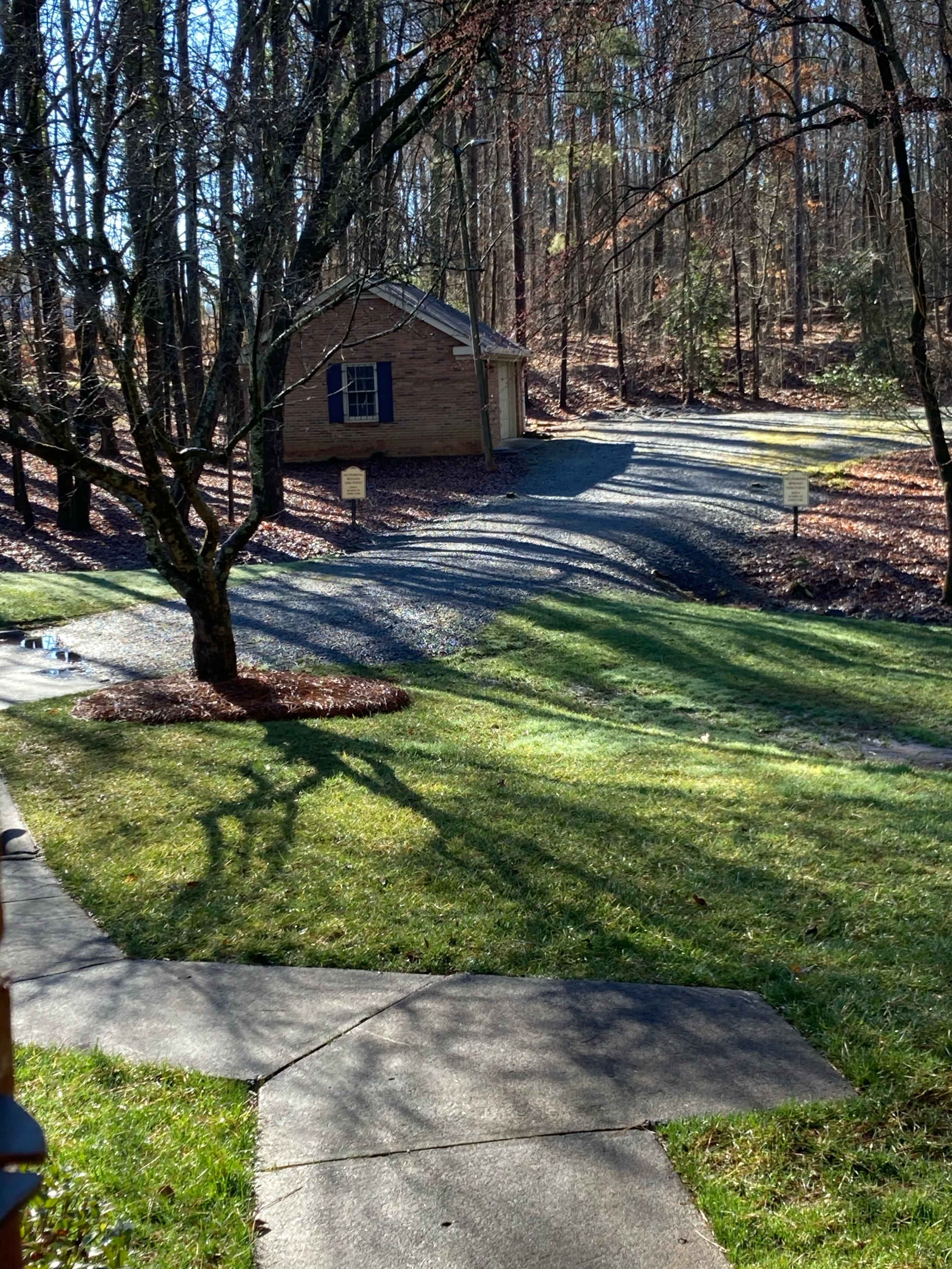 A small brick shed is situated at the end of a gravel driveway, surrounded by trees and a manicured lawn.