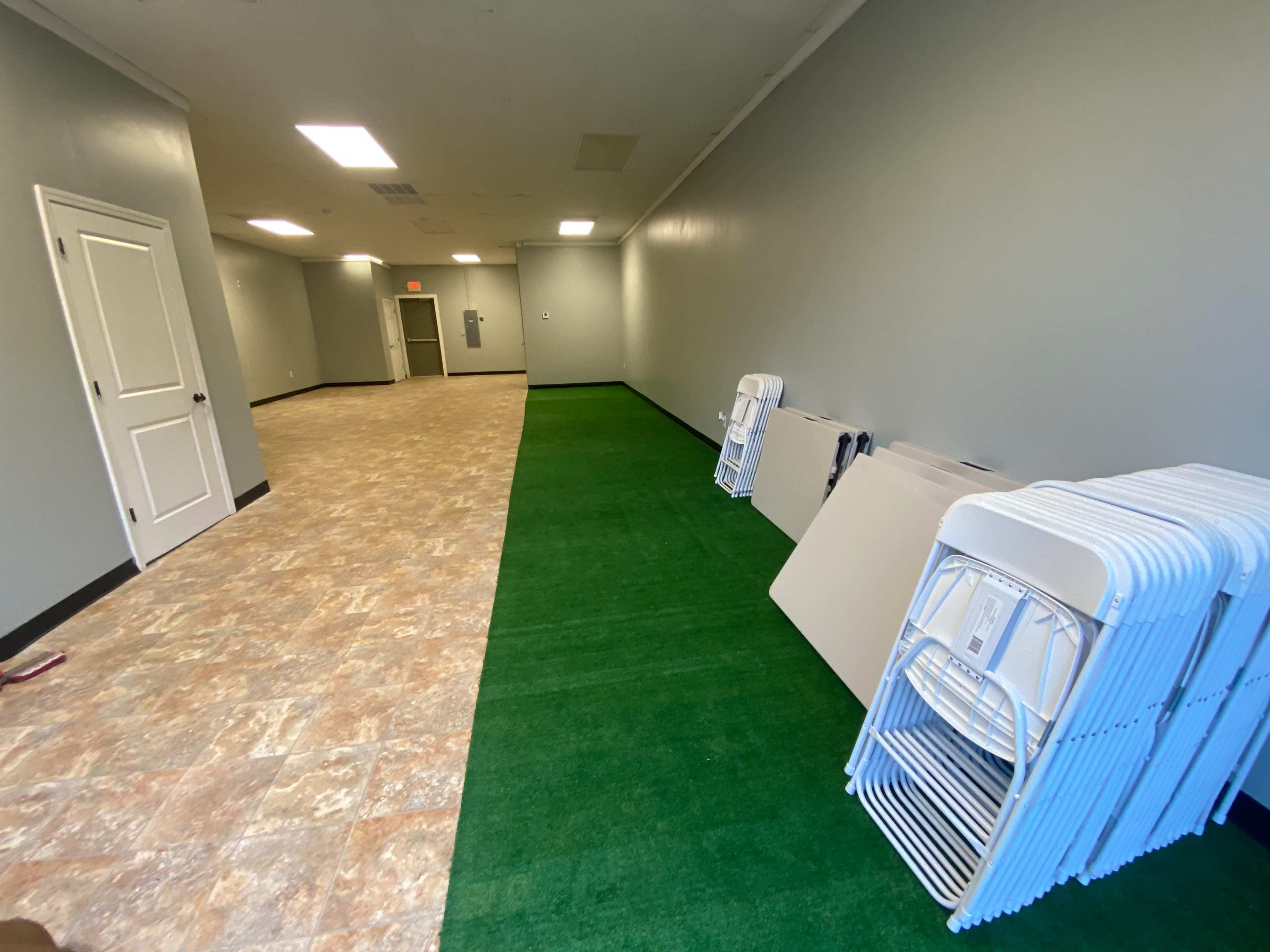 A narrow, well-lit hallway with green artificial grass on one side and stacked folding chairs and tables against the gray walls.