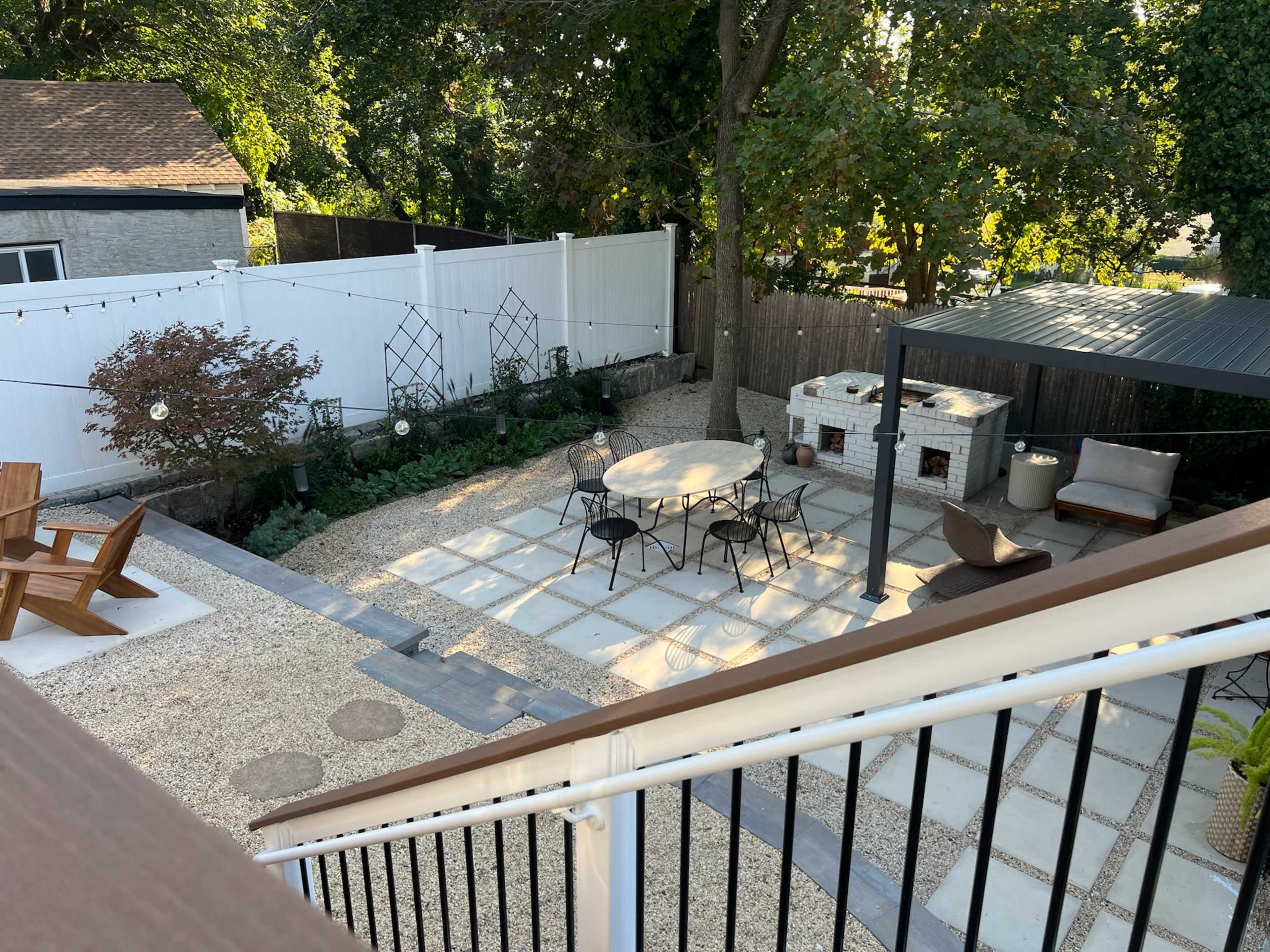 The image shows a landscaped backyard featuring a stone patio with a round table and chairs, surrounded by gravel, greenery, and an outdoor cooking area.