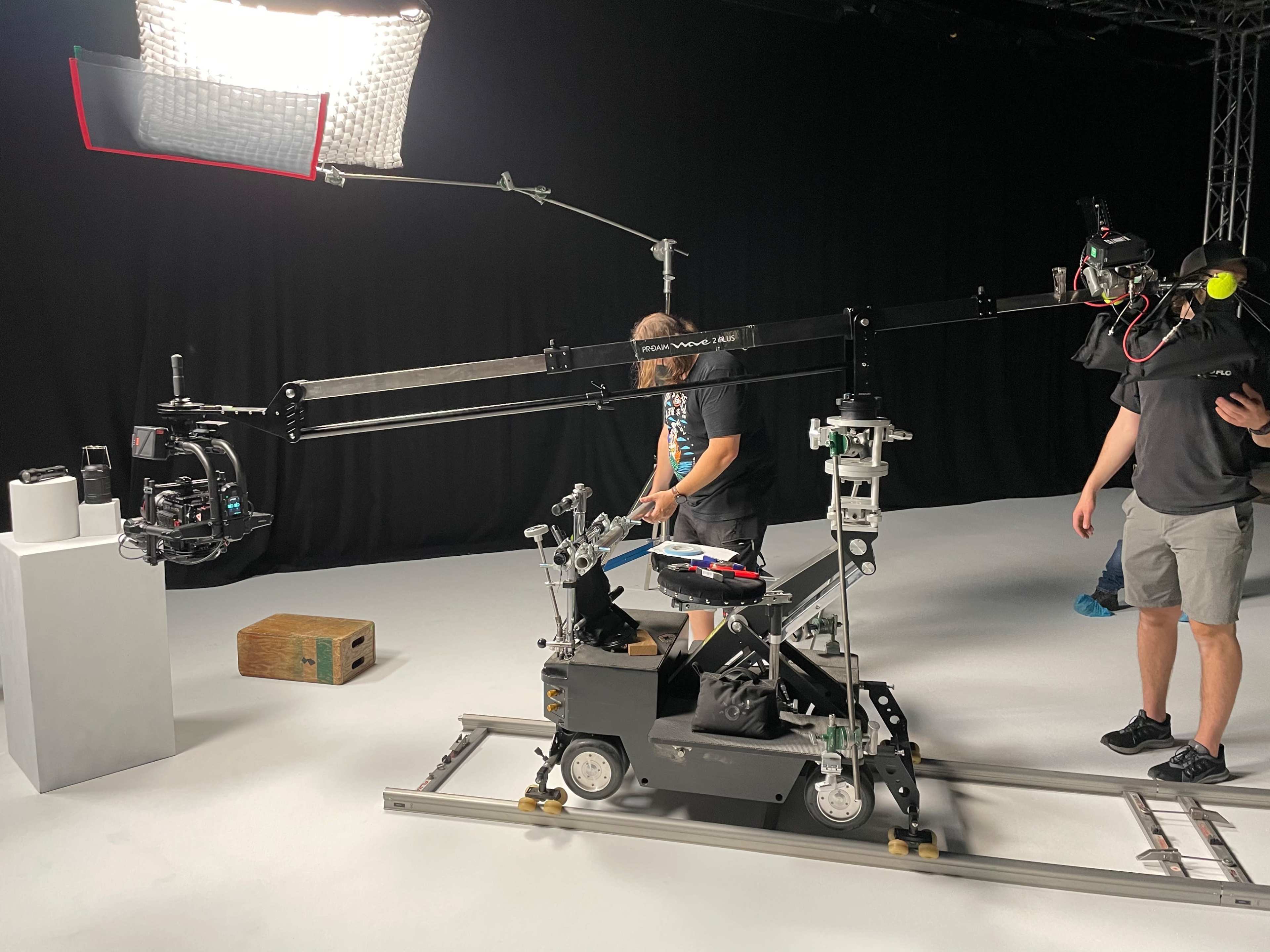 A camera setup is shown on a track with a crew member operating a tripod and a second crew member adjusting equipment in a studio setting.