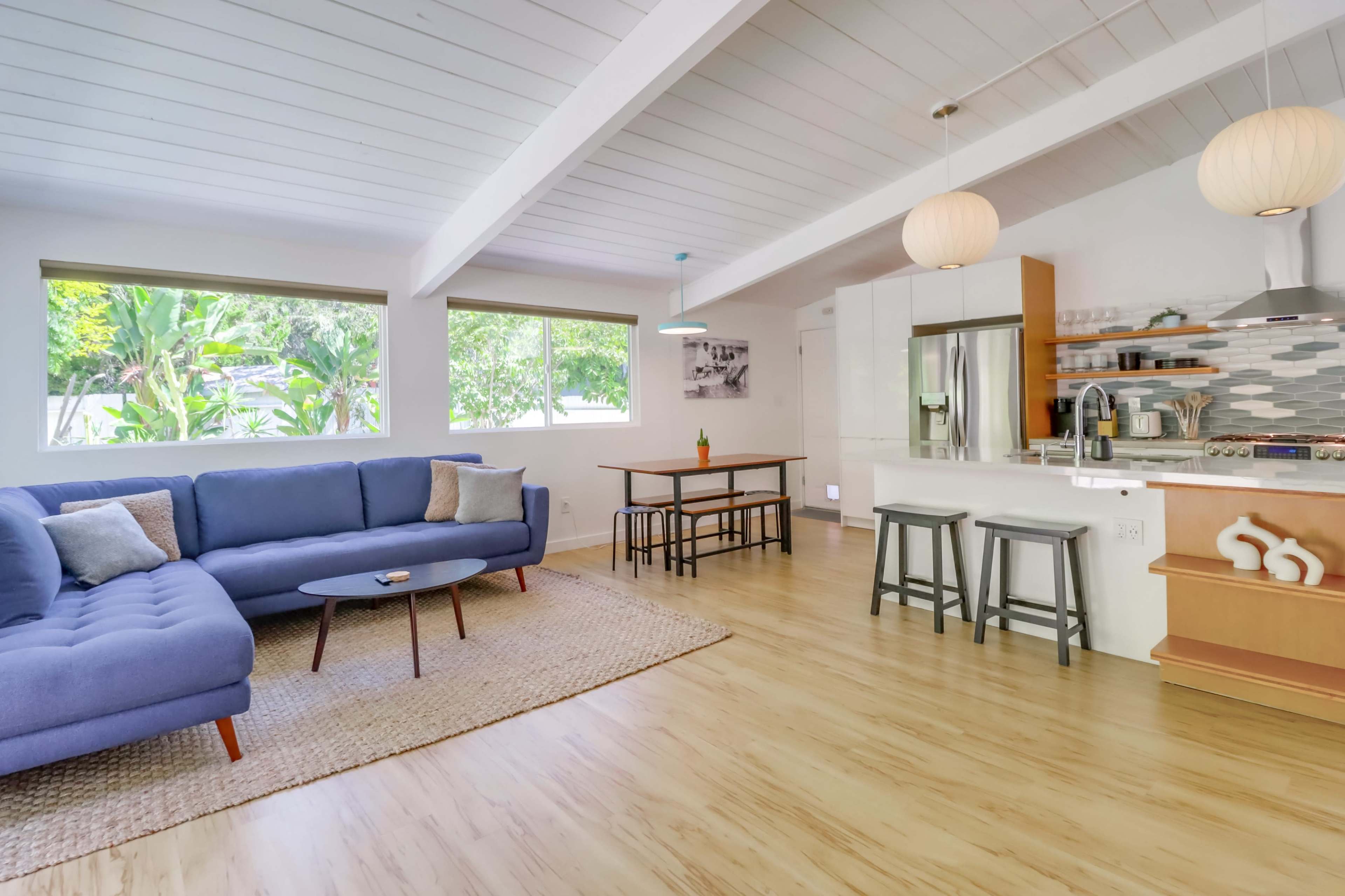 A modern, open-concept living area featuring a blue sectional sofa, a wooden dining table, and a kitchen with light-colored cabinetry and decorative elements.