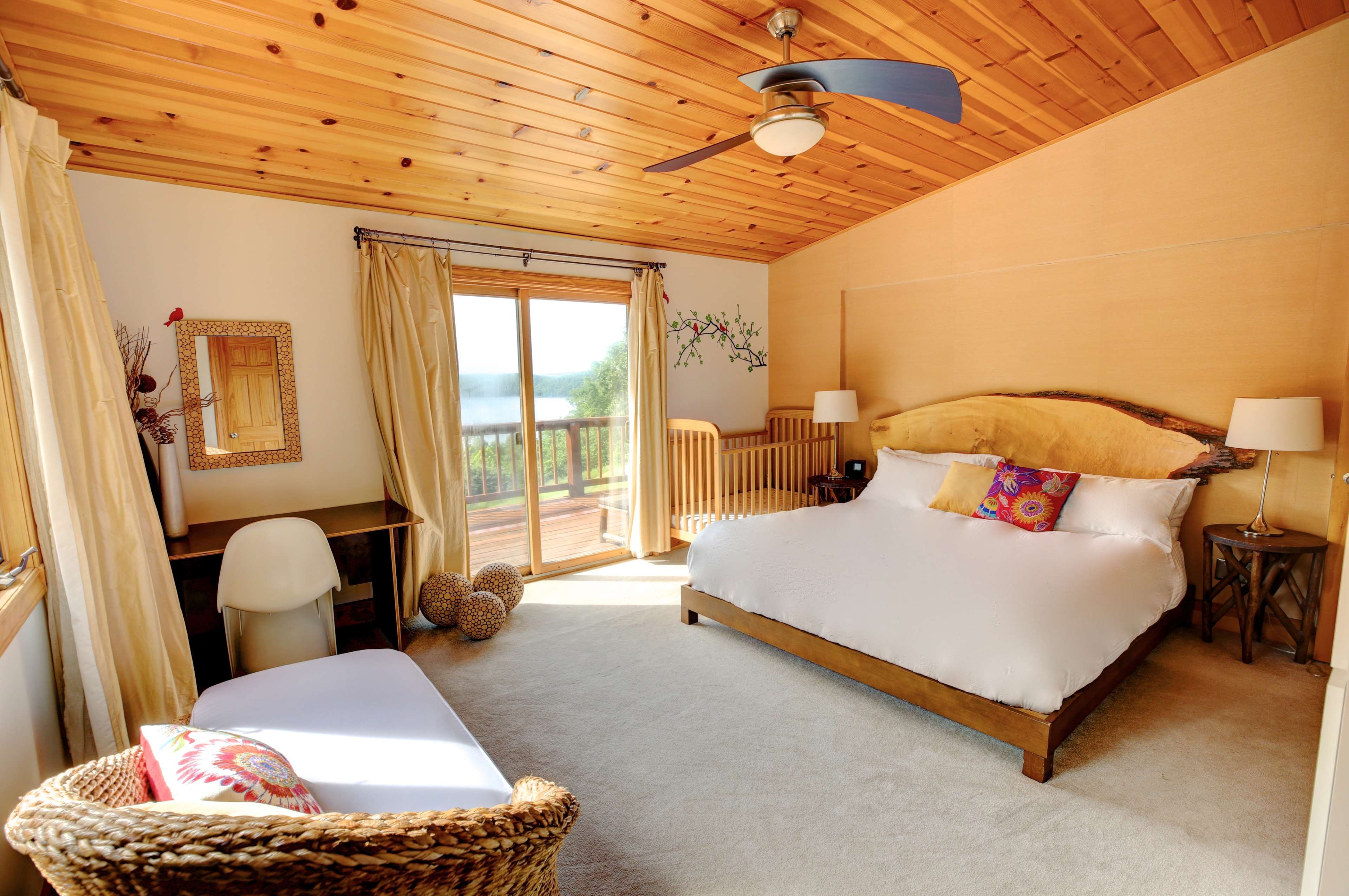 A well-lit bedroom featuring a king-sized bed, a wicker chair, a sliding glass door leading to a deck, and wooden ceiling panels.