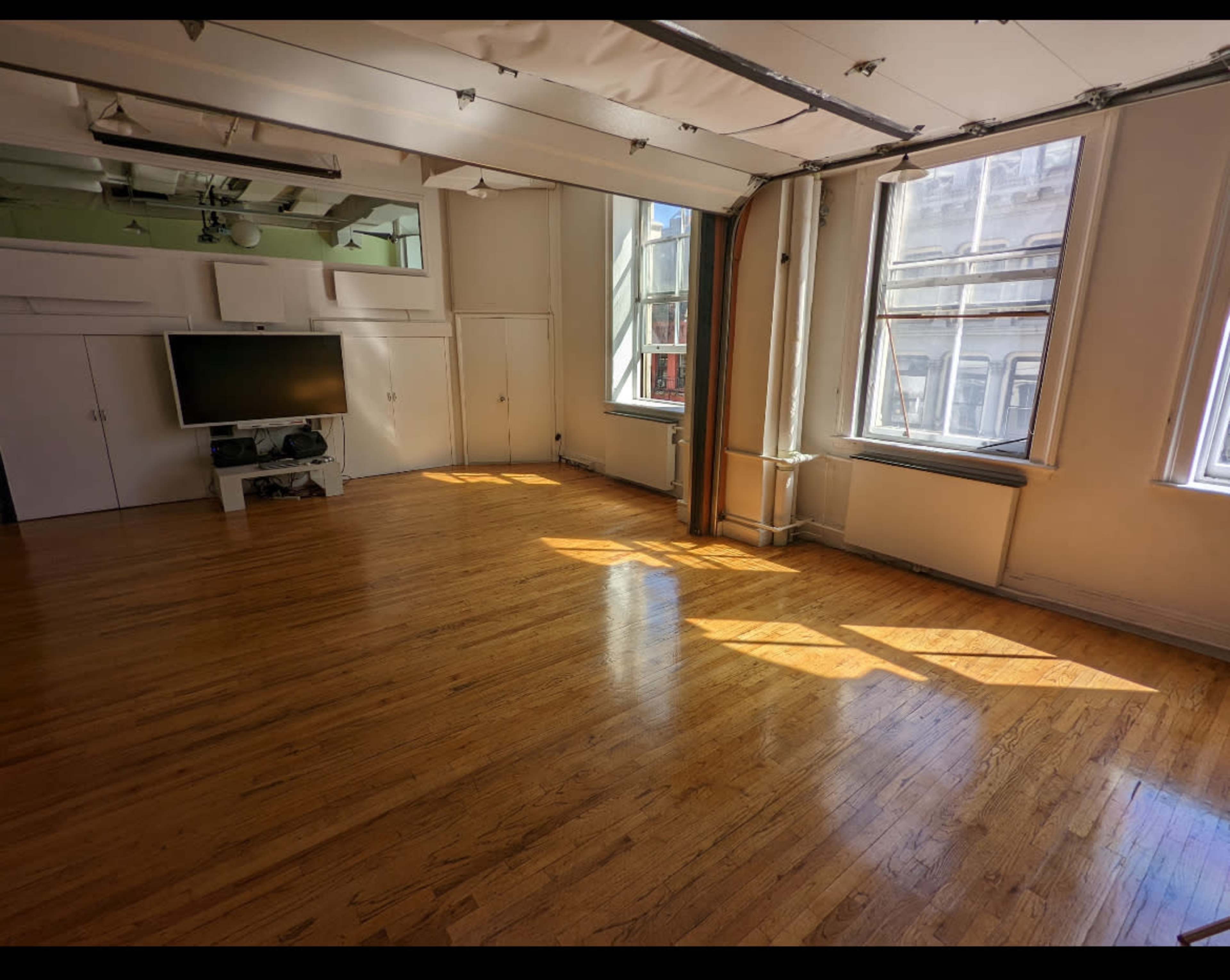A spacious room with hardwood floors, large windows allowing sunlight to stream in, and a television mounted on the wall.