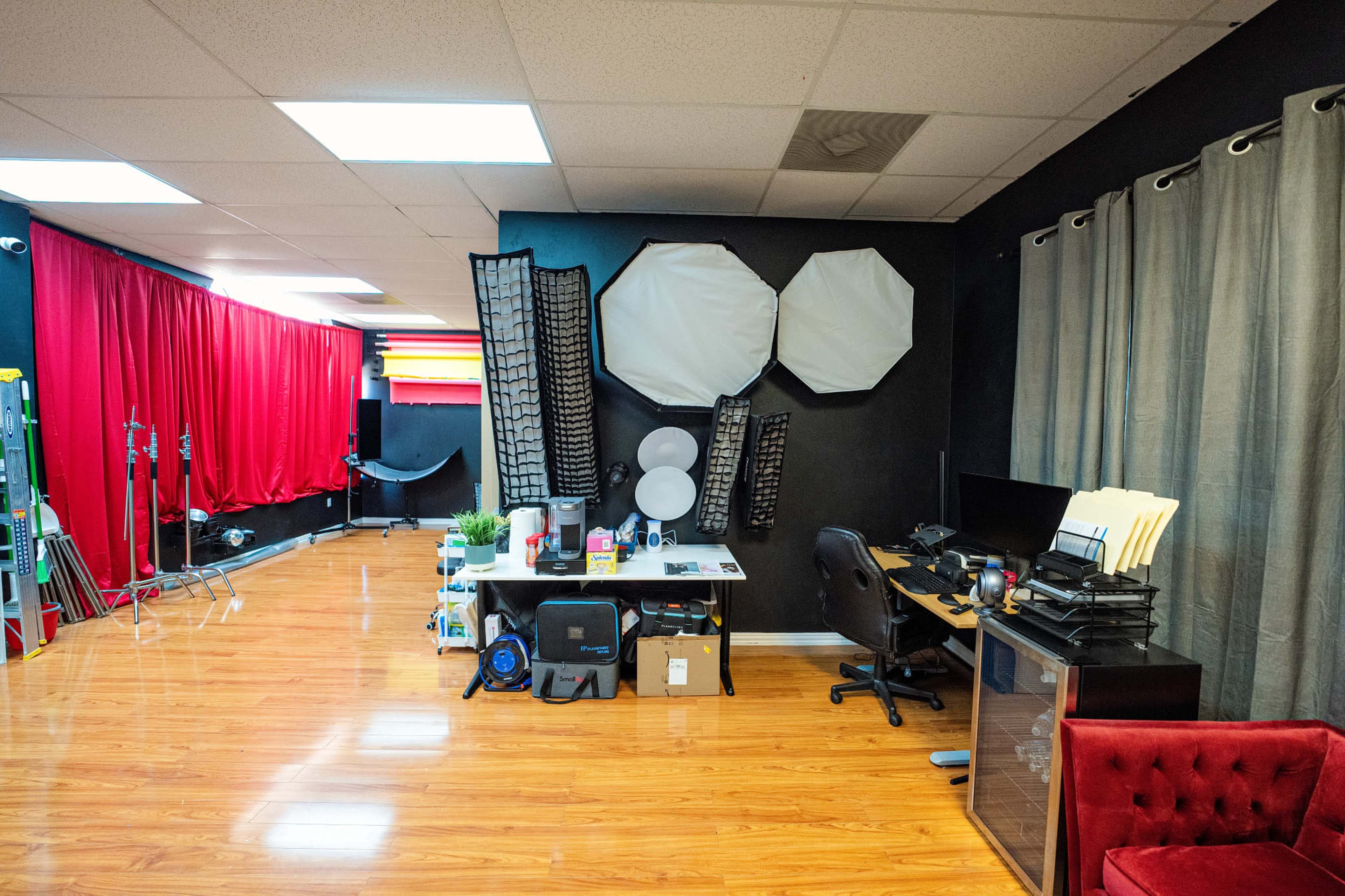 The image shows a studio space with red curtains on one side, various photography equipment on a table, and a desk with a computer and office chair against a black wall.