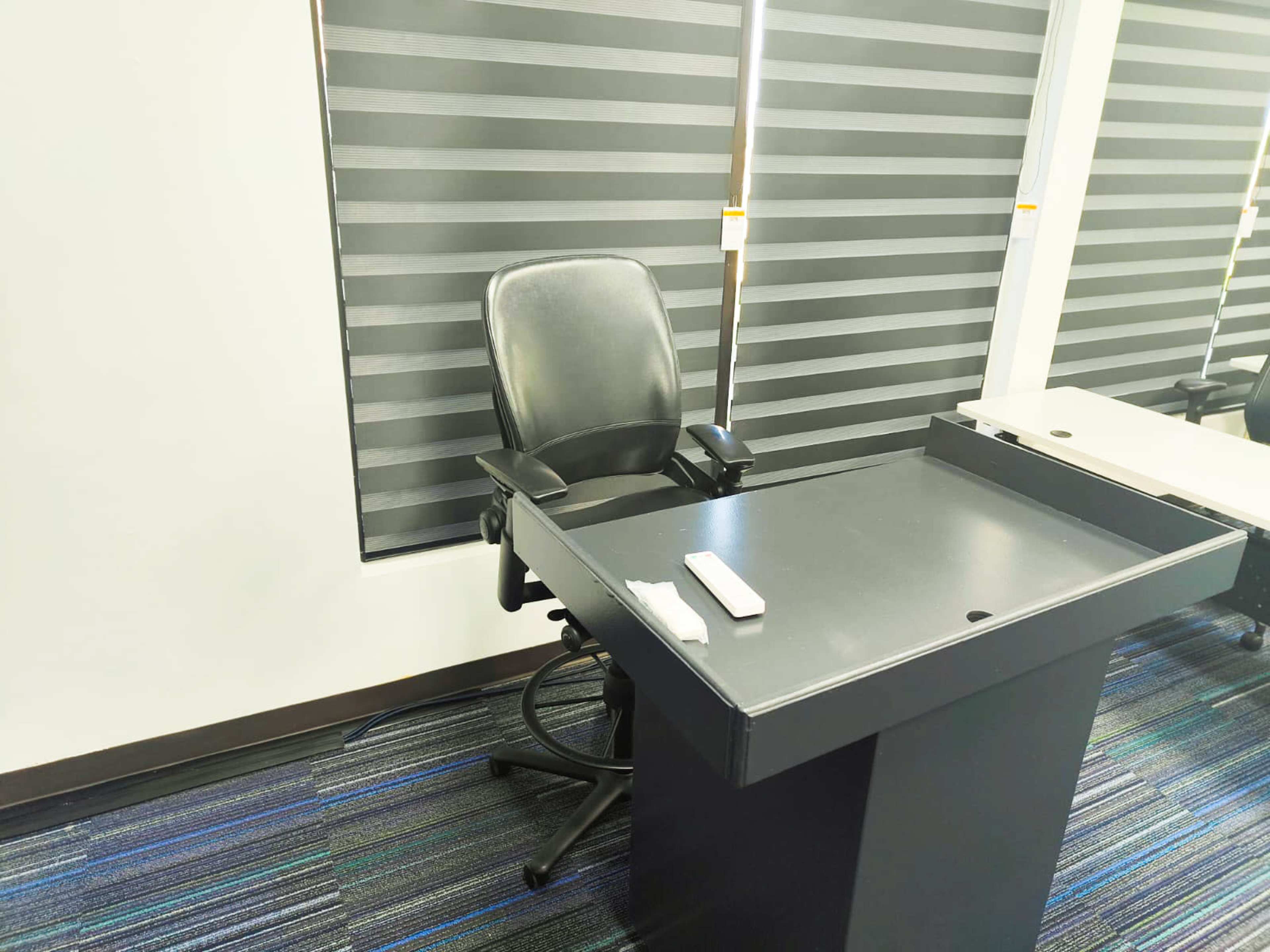 A black desk with a rolling chair beside it, located in a room with striped window blinds.