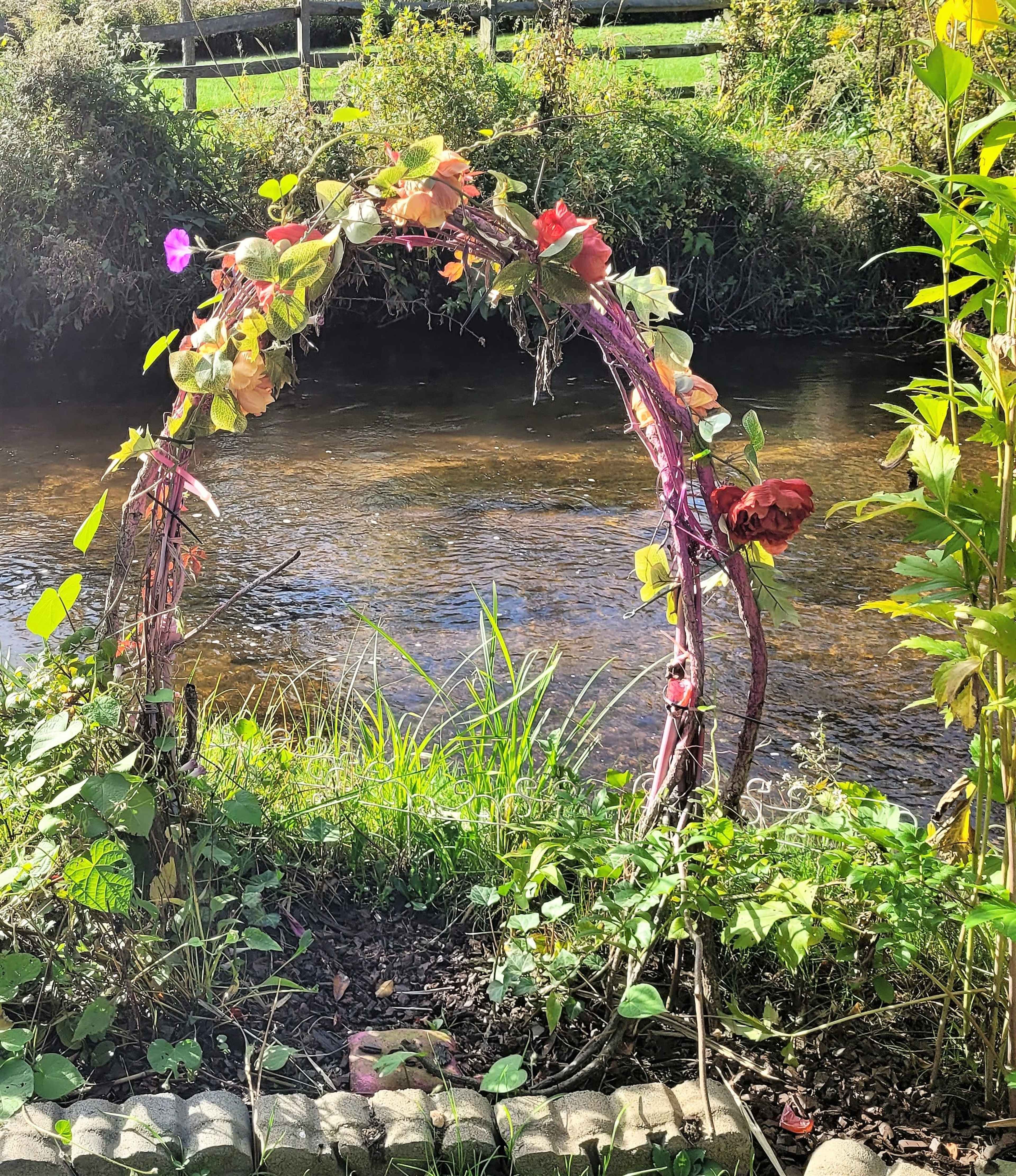 A vine-wrapped archway is positioned near a calm stream, surrounded by greenery.
