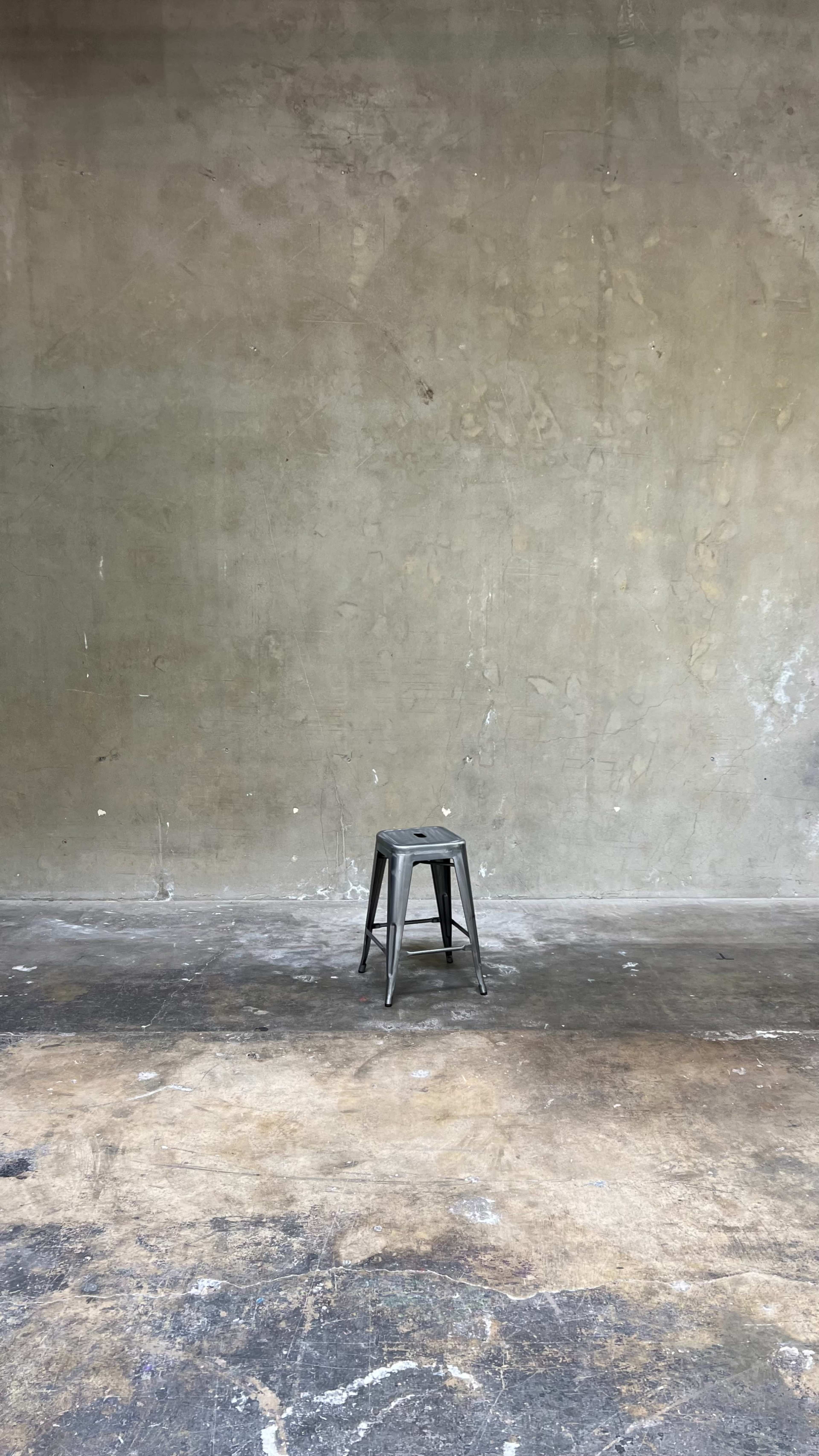 A lone metal stool stands on a bare, uneven concrete floor against a plain, textured wall.