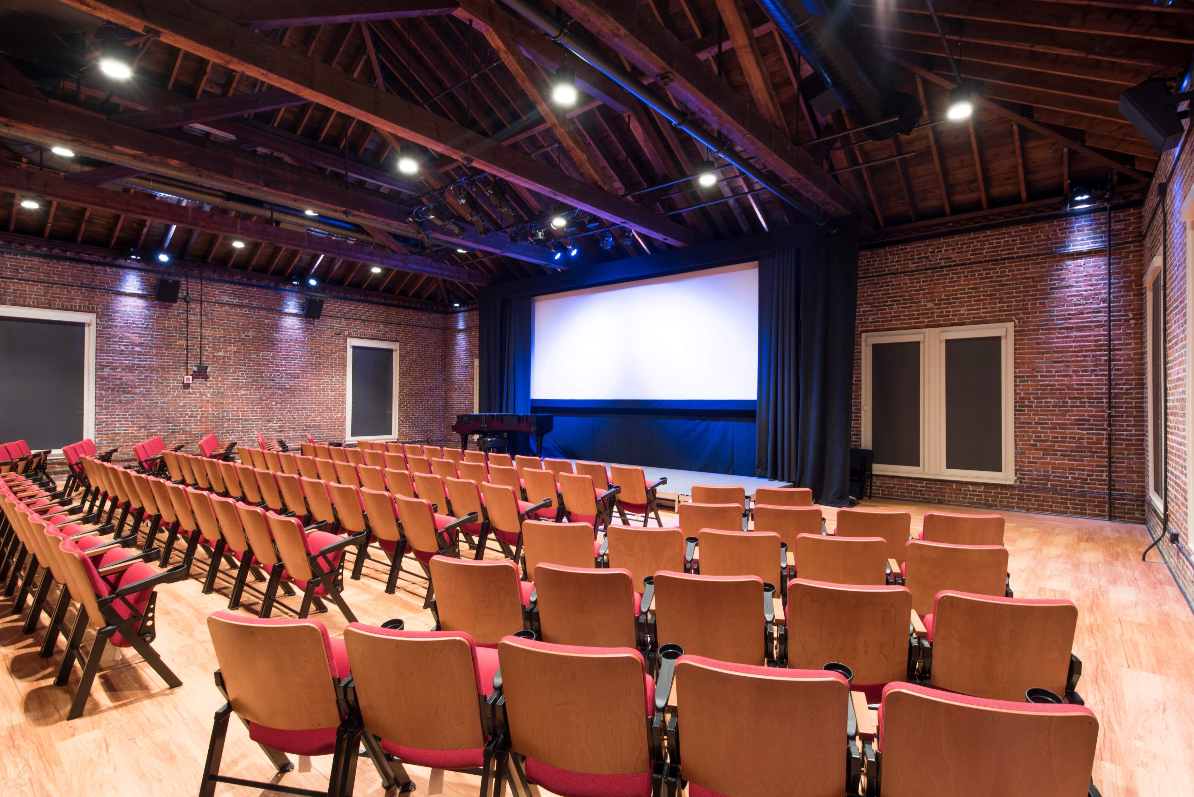 A small theater features rows of red cushioned seating facing a large projection screen, set against a backdrop of brick walls and wooden beams.