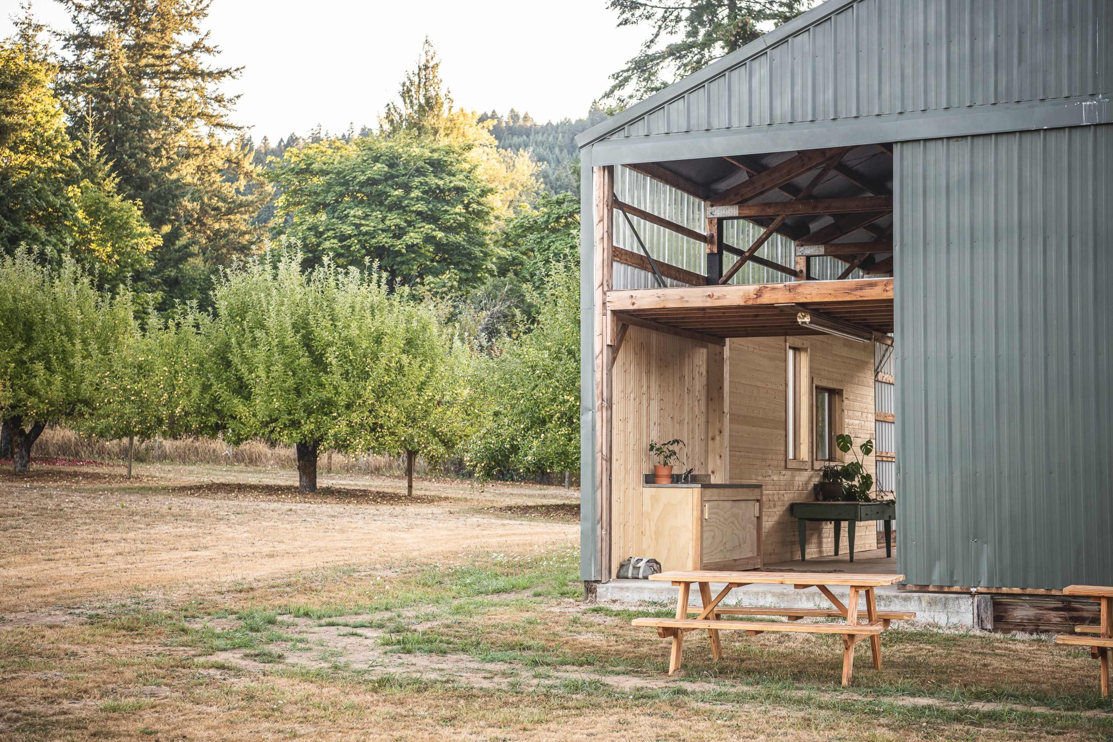 A metal-clad building with a wooden porch is set near an orchard of trees in a grassy area.
