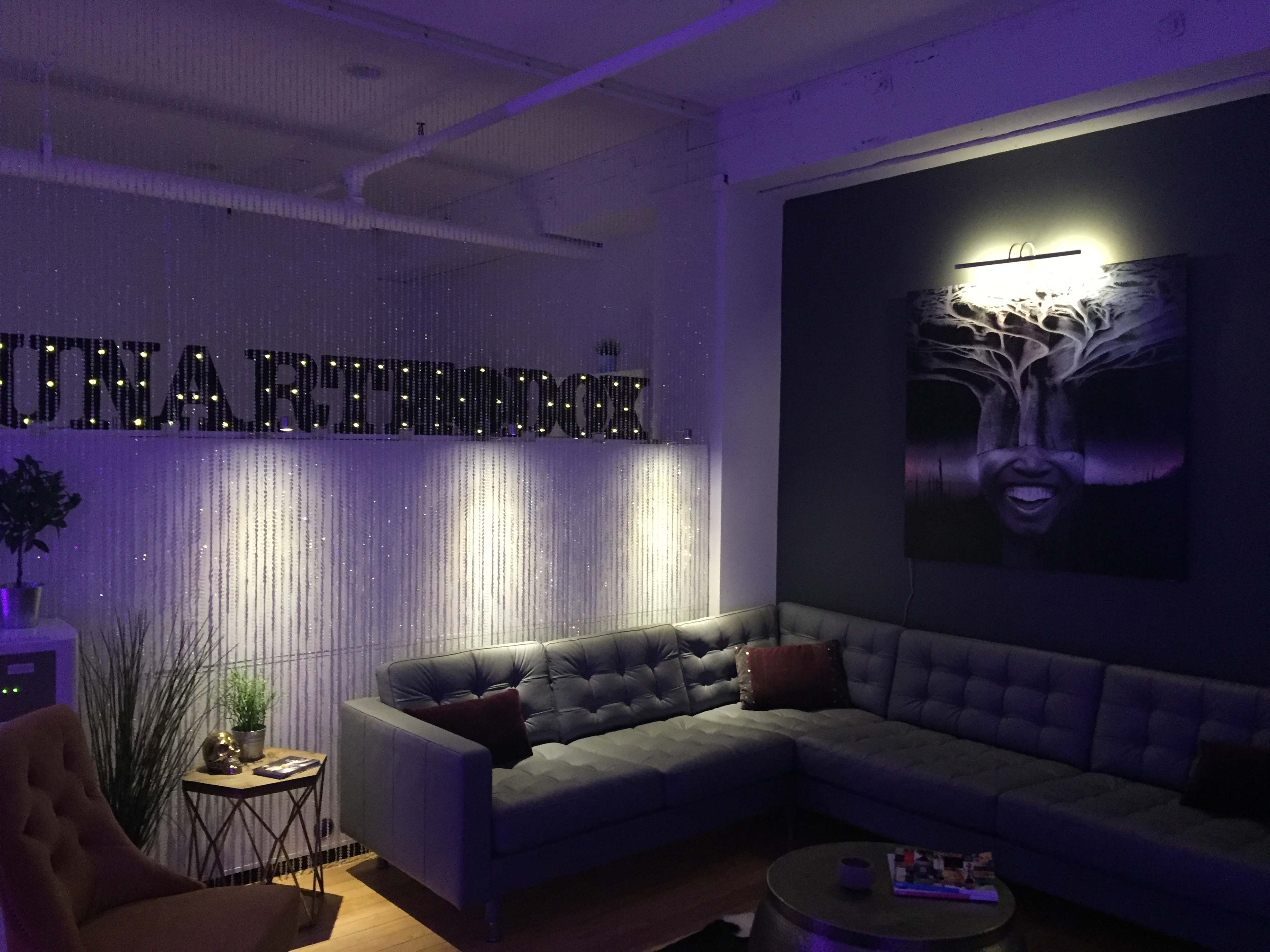 A modern lounge area with a gray sectional sofa, decorative wall art, and illuminated signage reading "LUNARTHOOD."