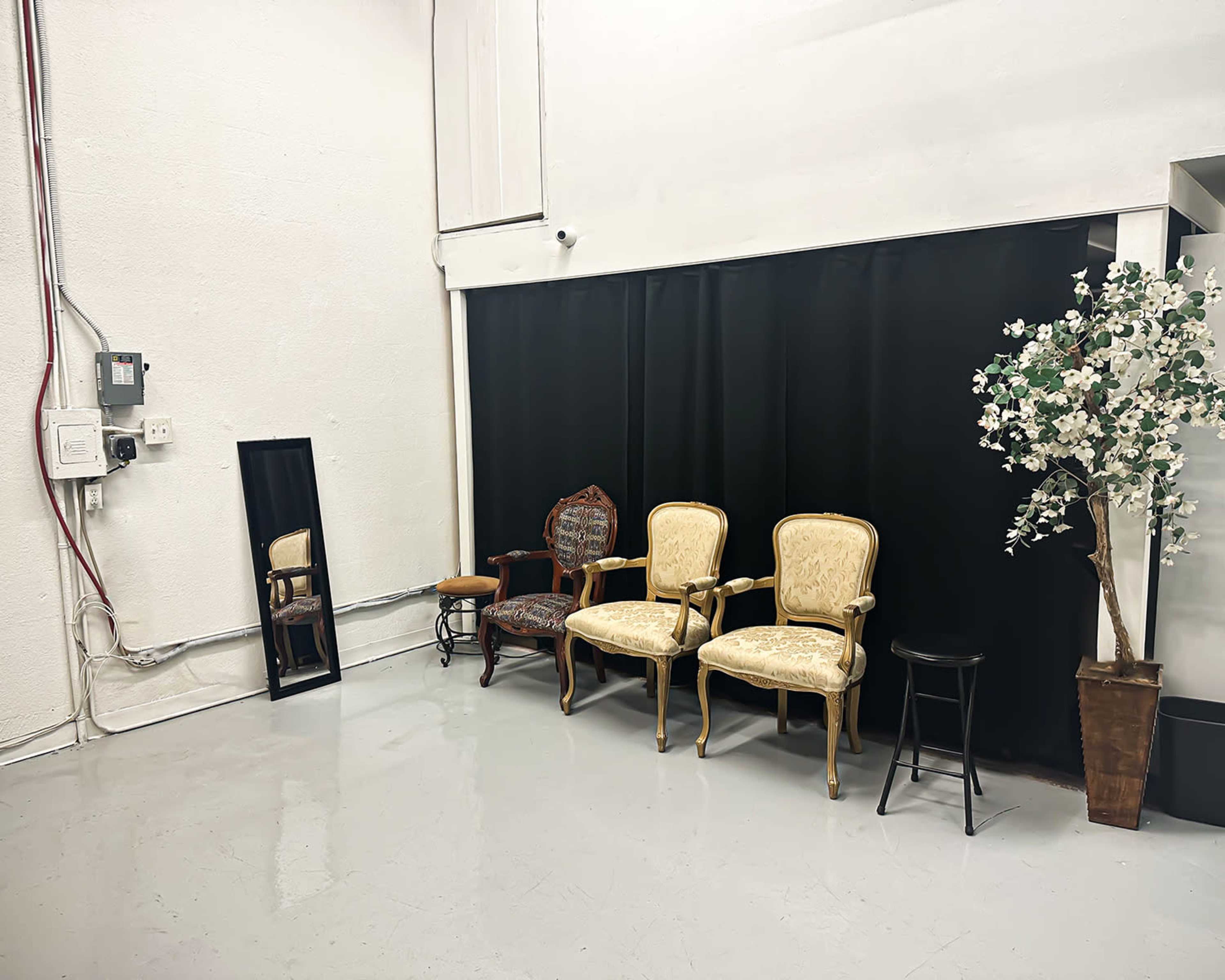 The scene shows a minimalist room featuring three chairs, a mirror, and a decorative plant near a black curtain.