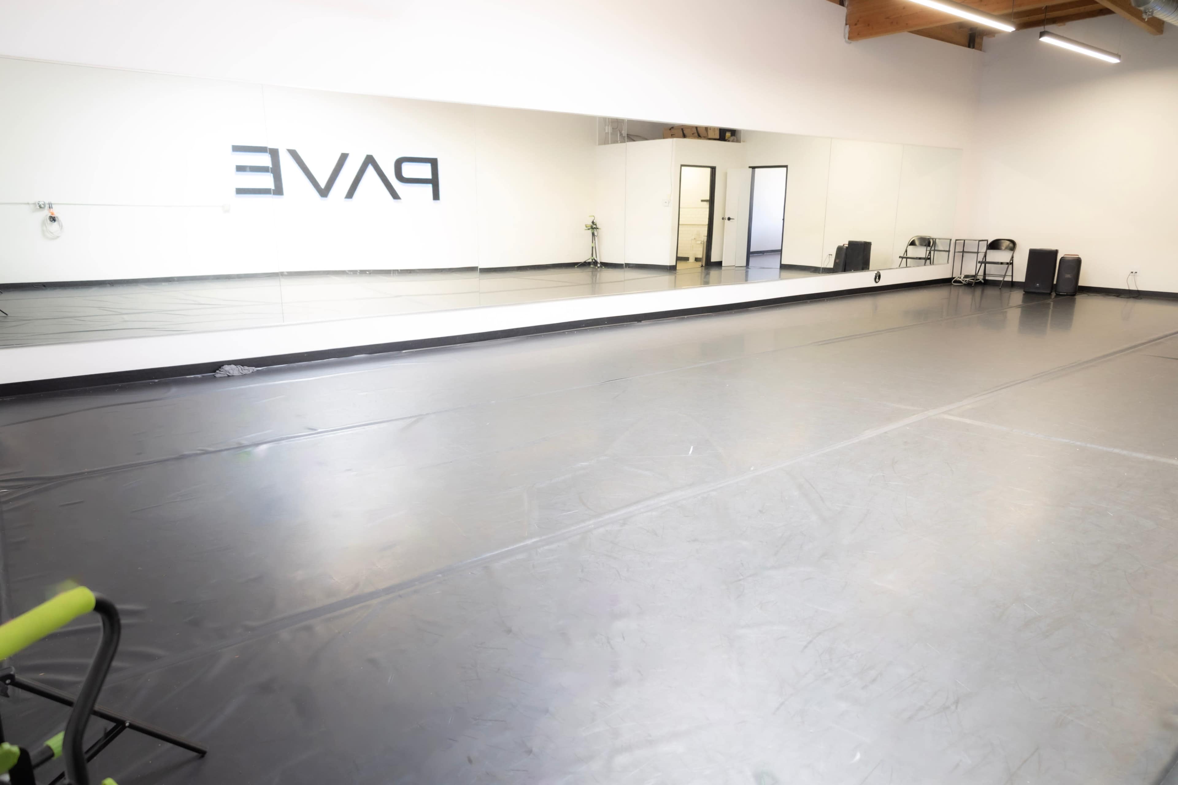 The image shows a spacious dance studio with a large mirror along one wall and a black rubberized flooring surface.