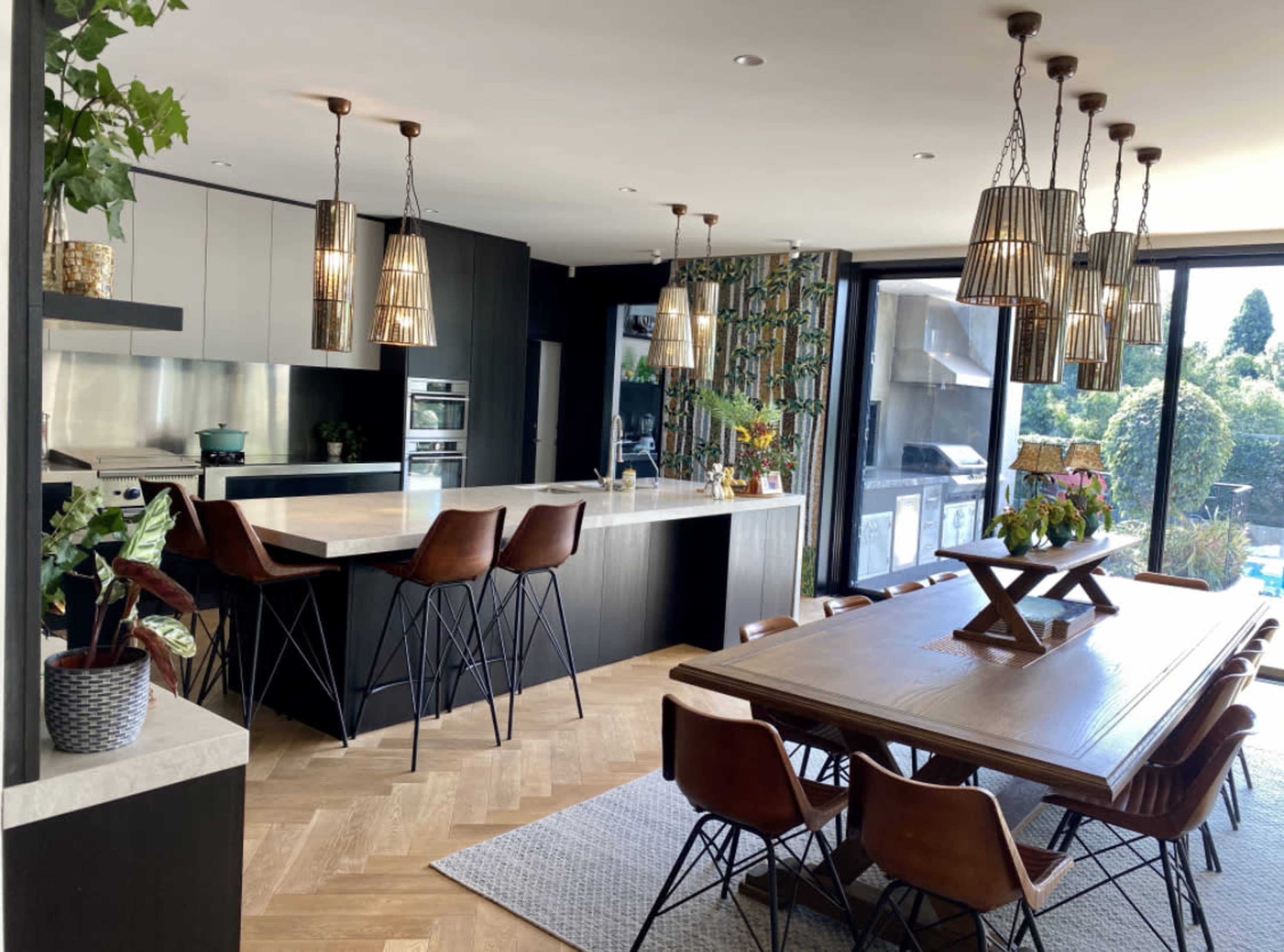 The image shows a modern kitchen and dining area with a large wooden table, sleek cabinetry, and multiple pendant lights hanging above the island.